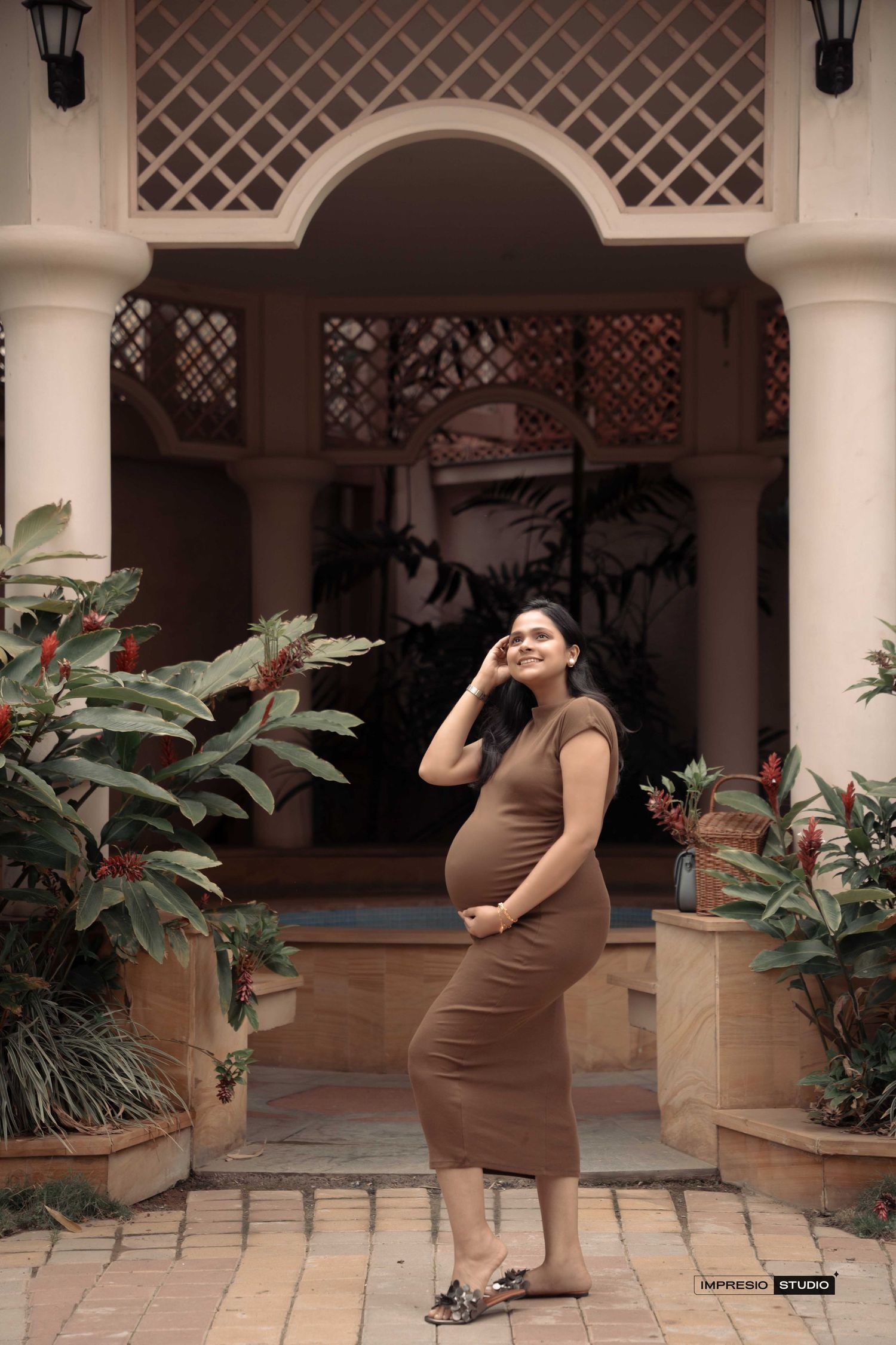 Pregnant woman in brown dress posing outdoors near tropical plants and elegant arched building entrance.