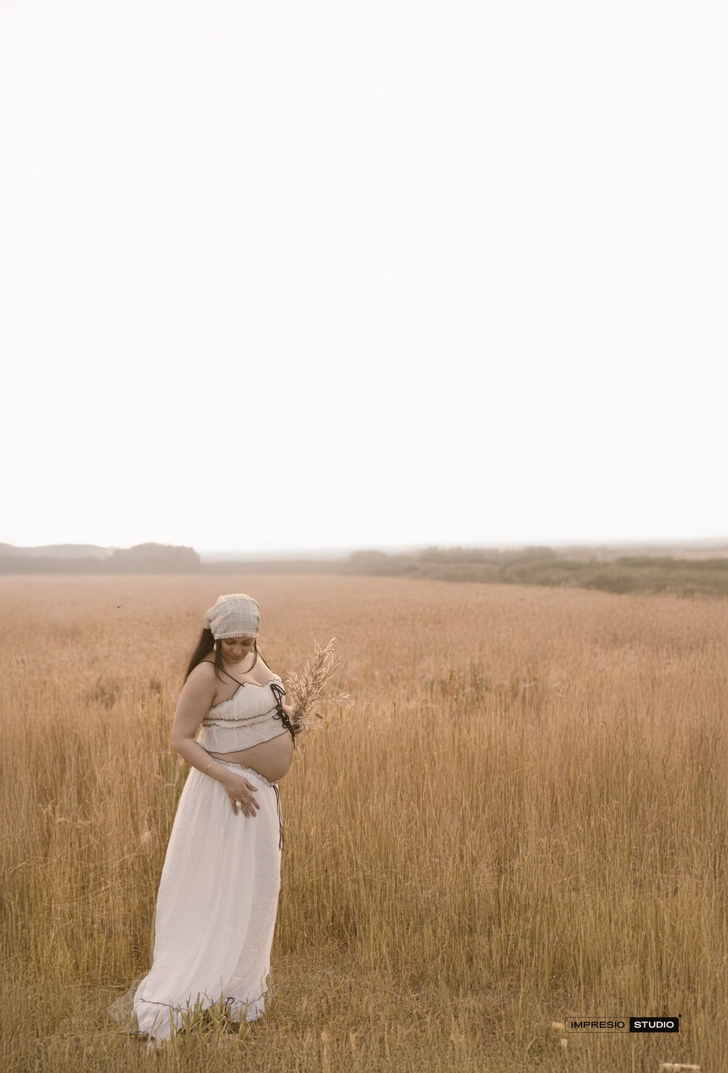 Pregnant woman in white dress and hat stands in golden field holding wildflowers during a serene maternity photo session.