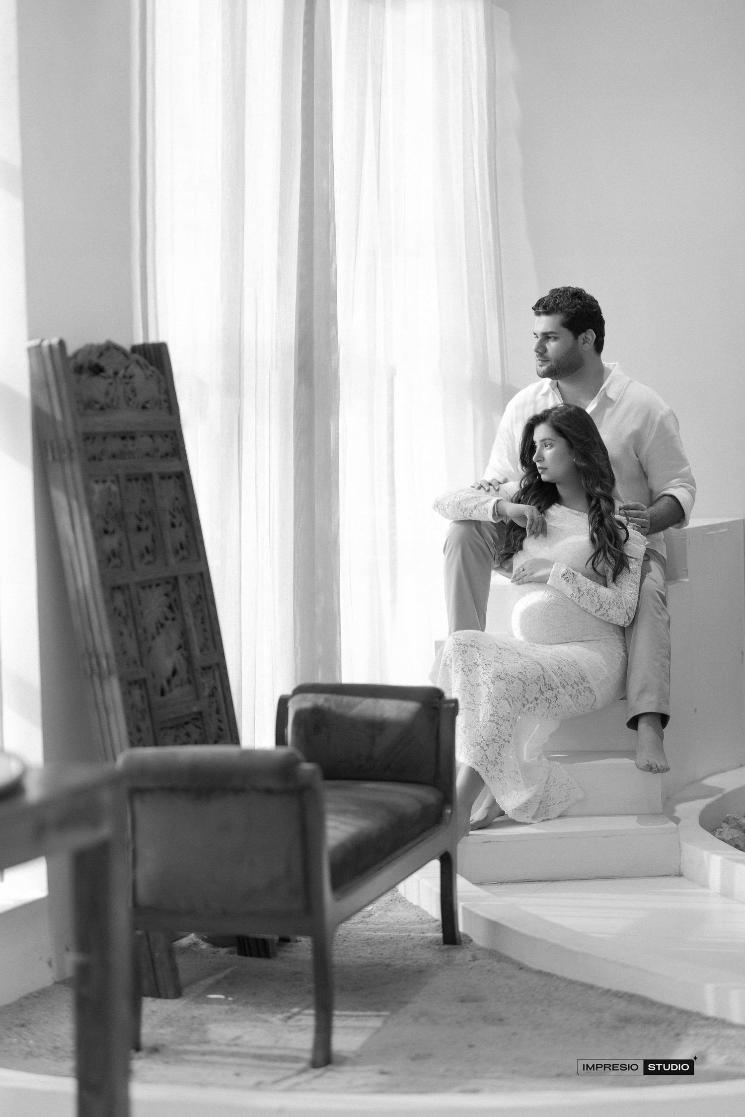 Black and white maternity photo of a couple in a bright room, woman in lace dress seated as man stands beside her.