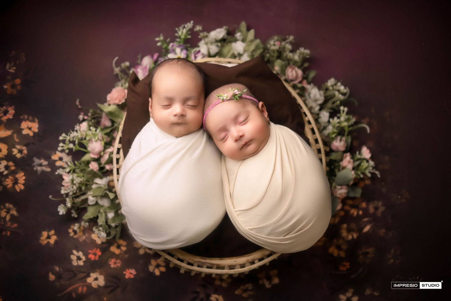 Two newborn twins swaddled in white and cream wraps, sleeping peacefully in a floral basket on a dark background.