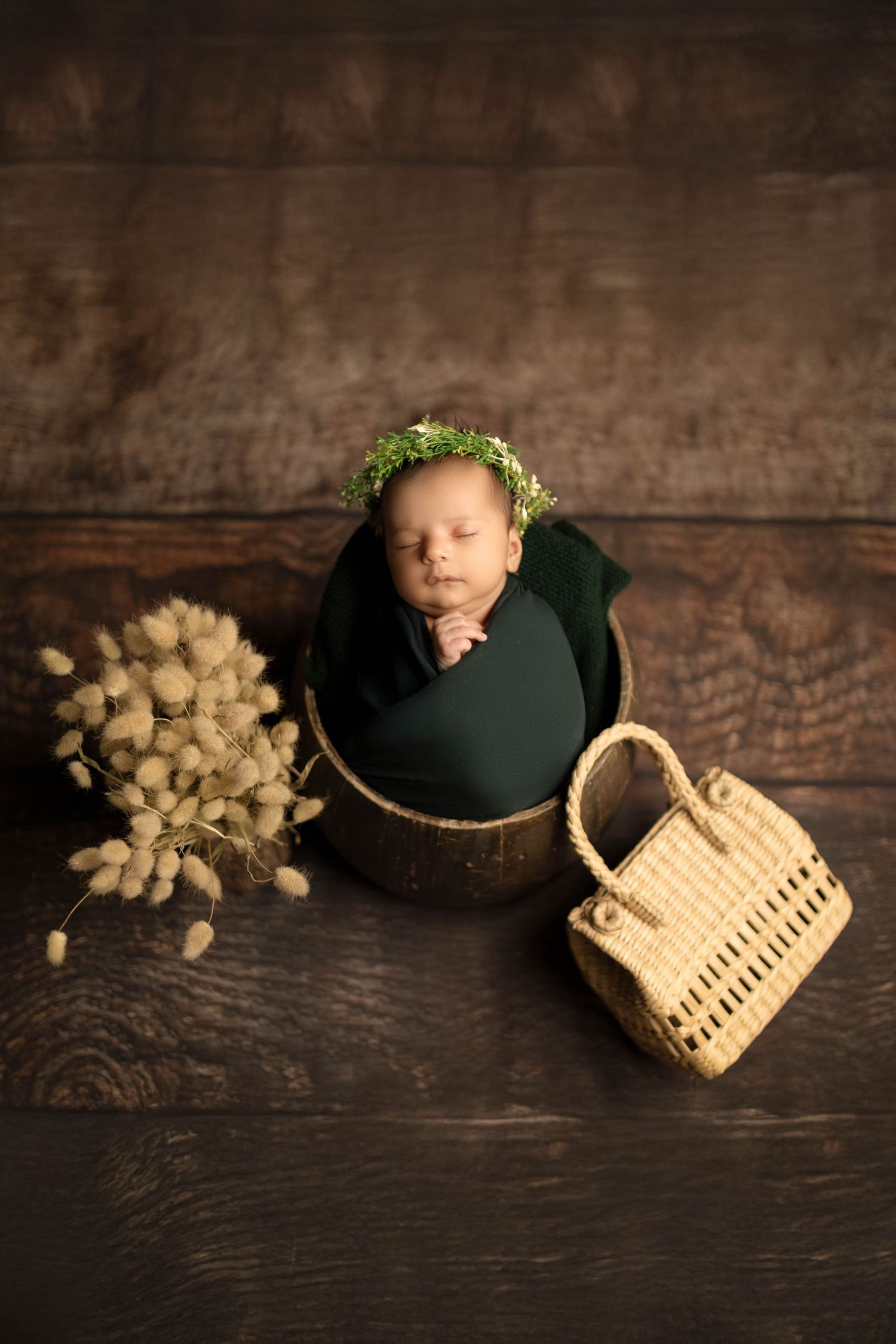 Newborn baby sleeping in a wooden bucket wrapped in green cloth, wearing a floral crown, beside dried flowers and a wicker basket.