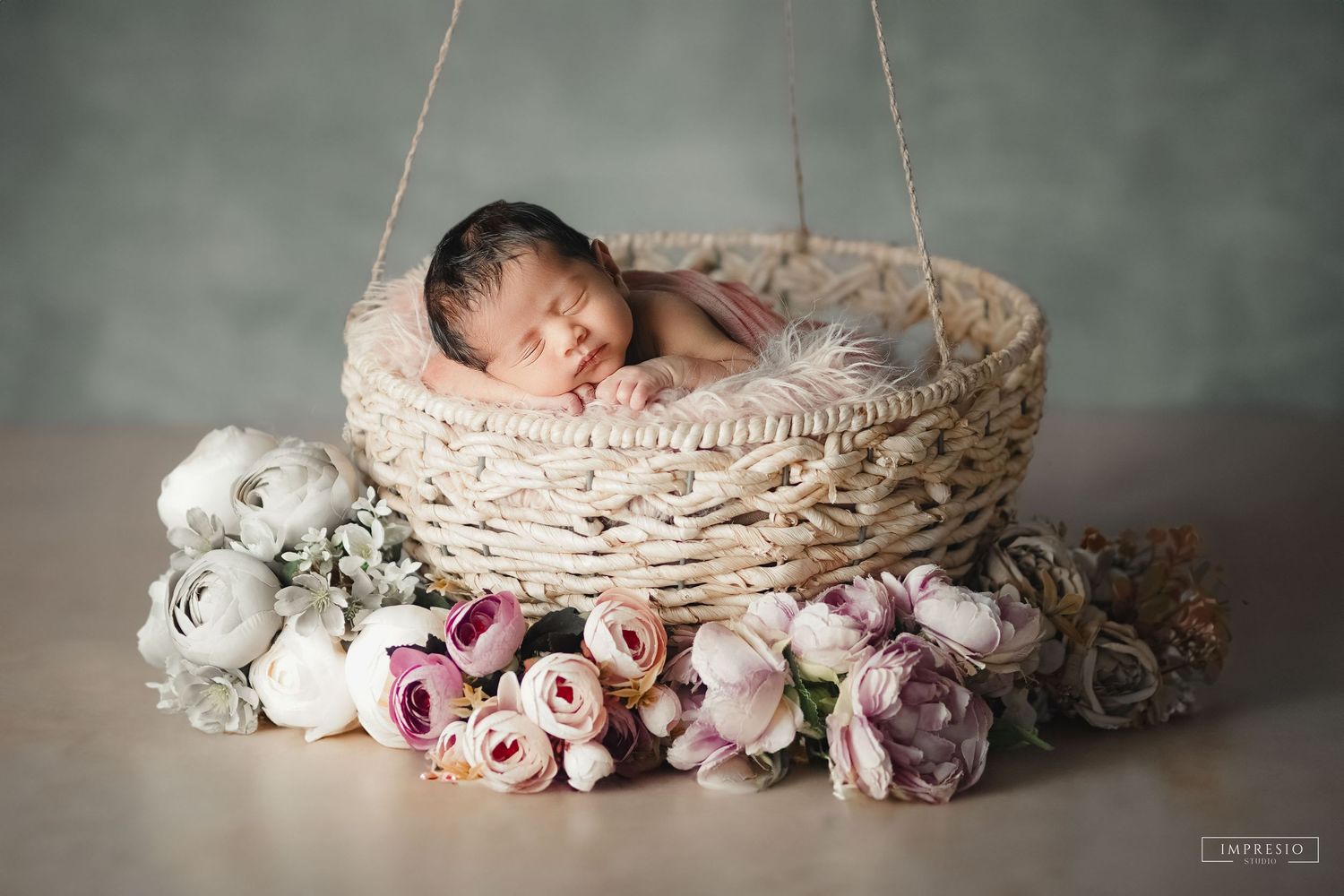 Newborn baby sleeping peacefully in a woven basket surrounded by soft pink and white flowers.