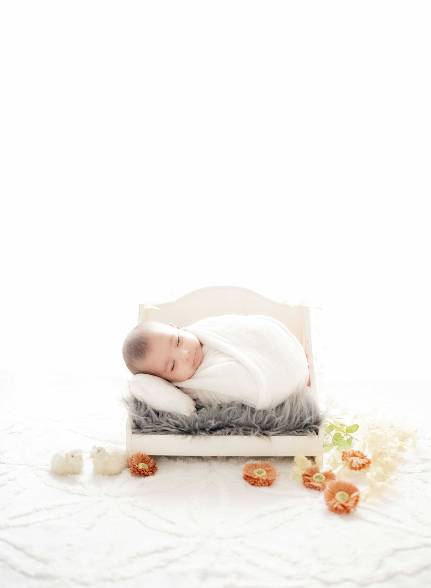 Sleeping newborn baby wrapped in white swaddle on a tiny wooden bed with gray fur and orange flowers.