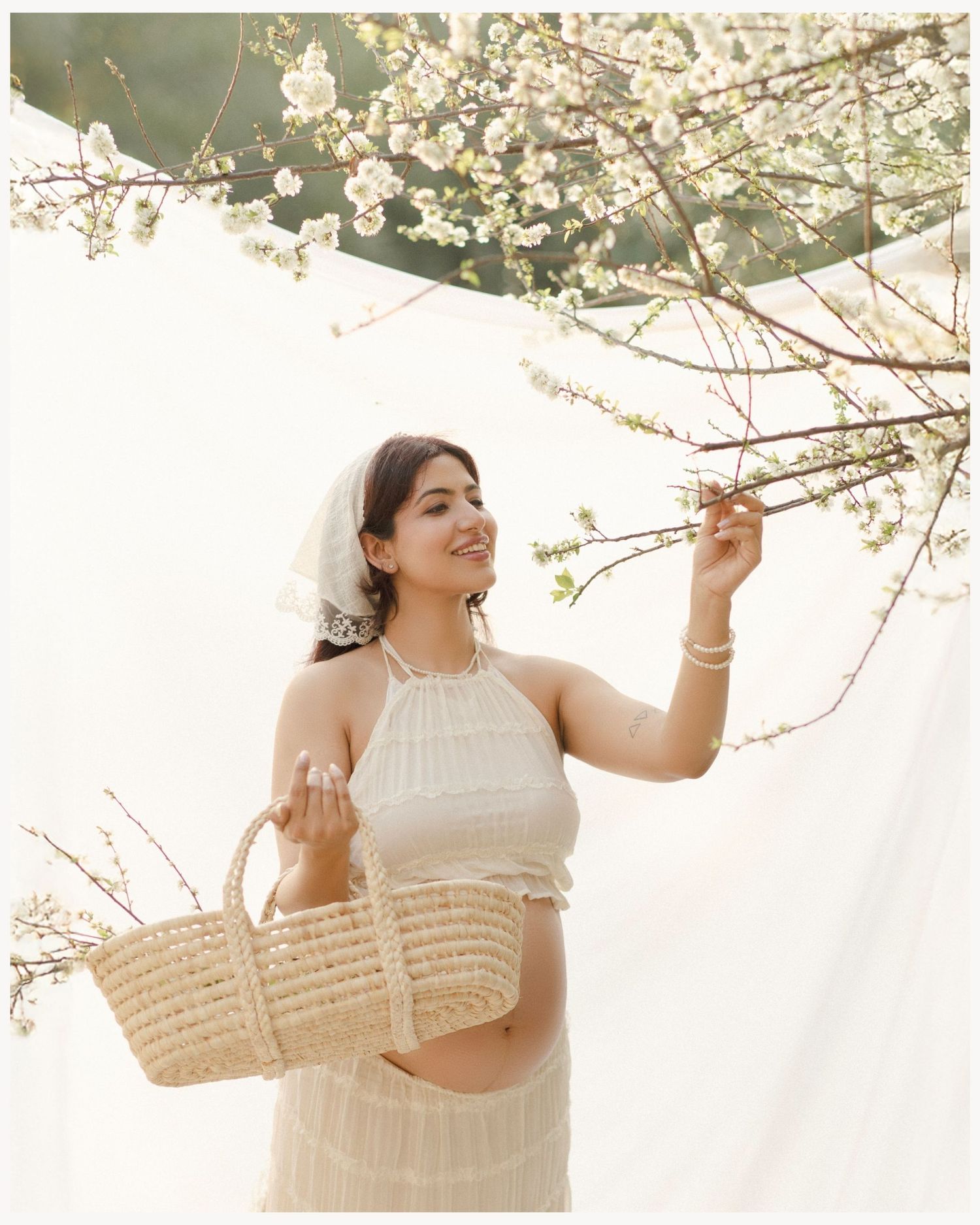 Smiling pregnant woman in white halter crop top holding wicker basket under blooming white cherry blossom tree.