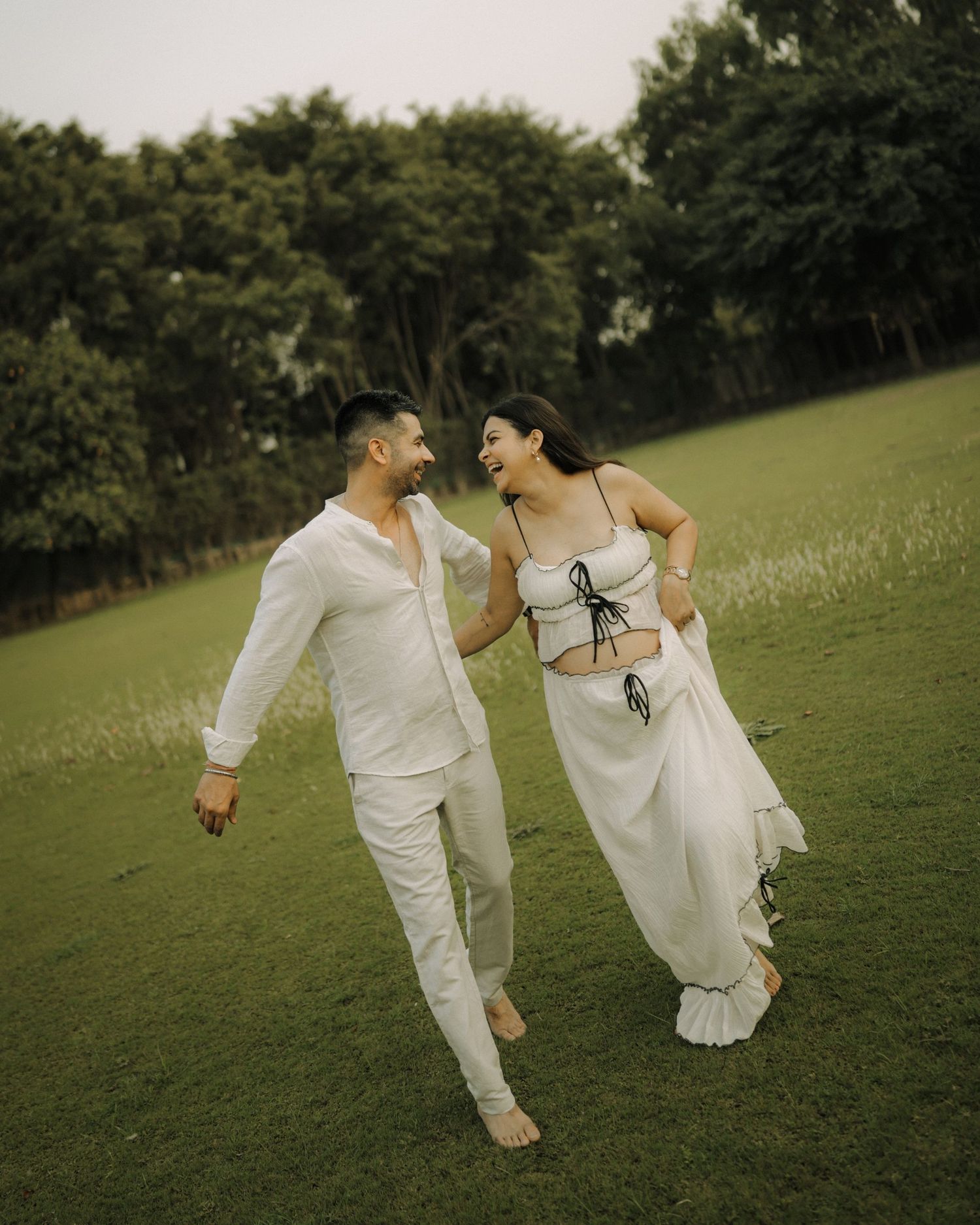 A couple in white outfits walk hand-in-hand laughing on a green lawn with lush trees in the background.