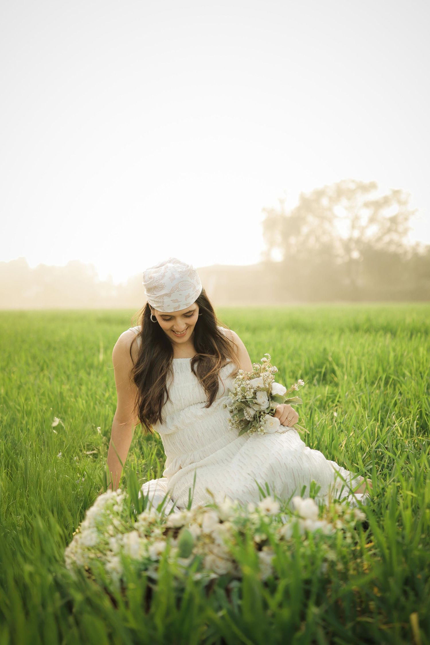 Bride in white dress and hat sits in lush green field holding a bouquet of white flowers in golden sunlight.