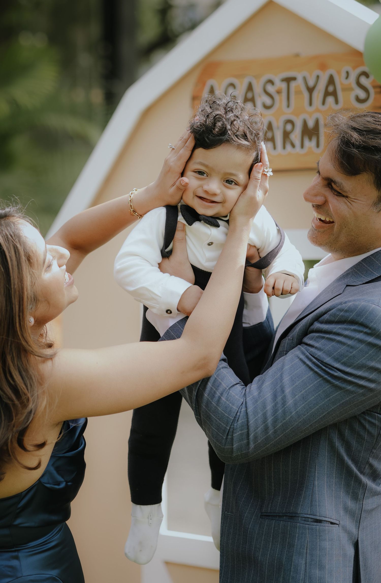 Parents lovingly hold their smiling baby boy in formal attire at Agastya's Farm outdoor celebration.