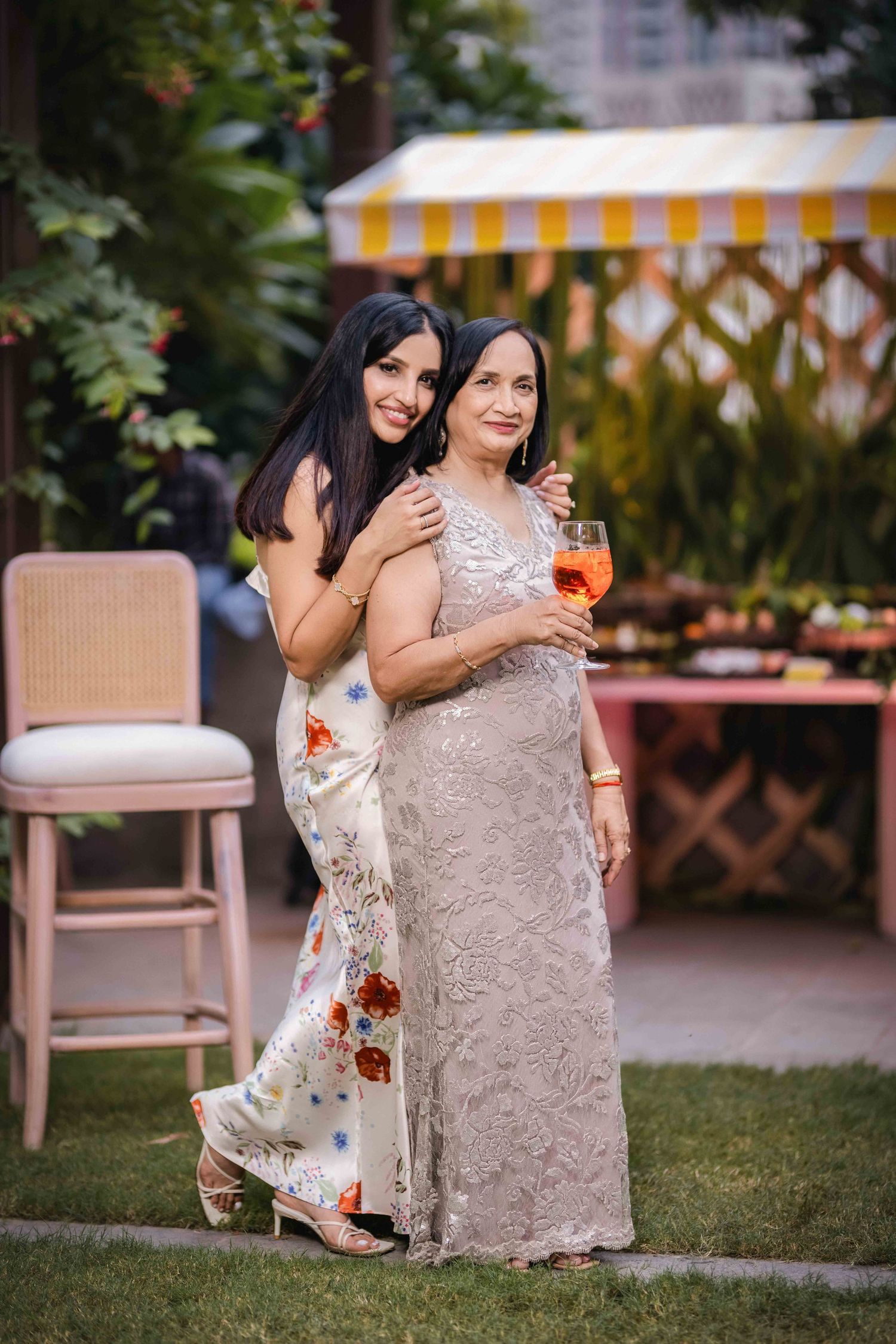 Two elegant women in formal gowns pose together at an outdoor garden party, one holding a cocktail drink.