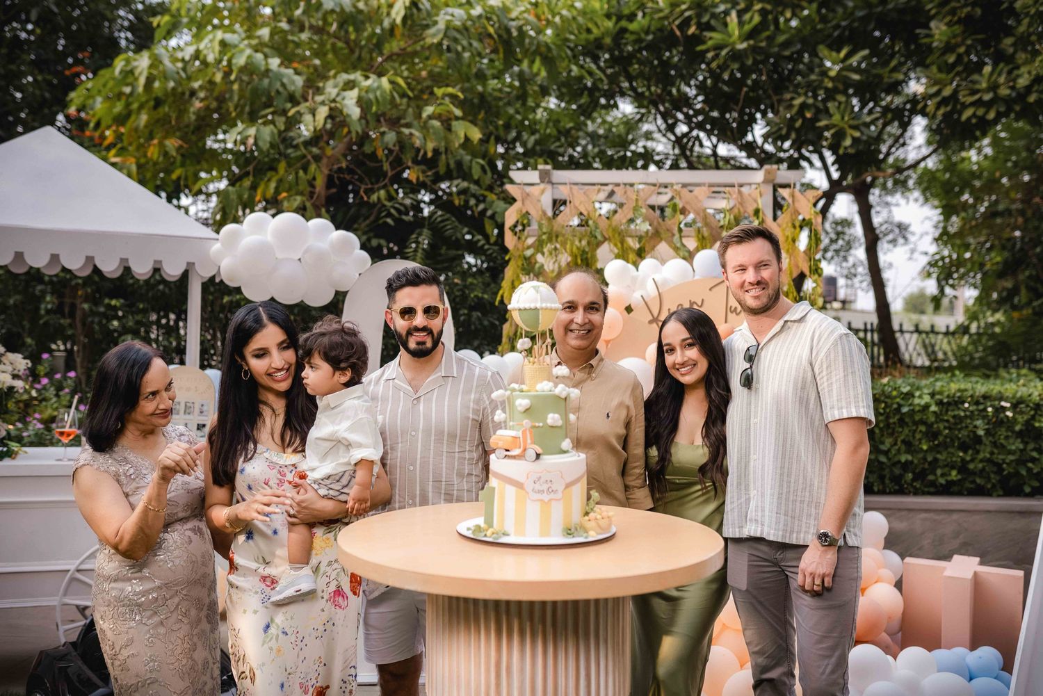 Group of six adults and a toddler celebrating a birthday outdoors with a decorated cake on a pedestal table.