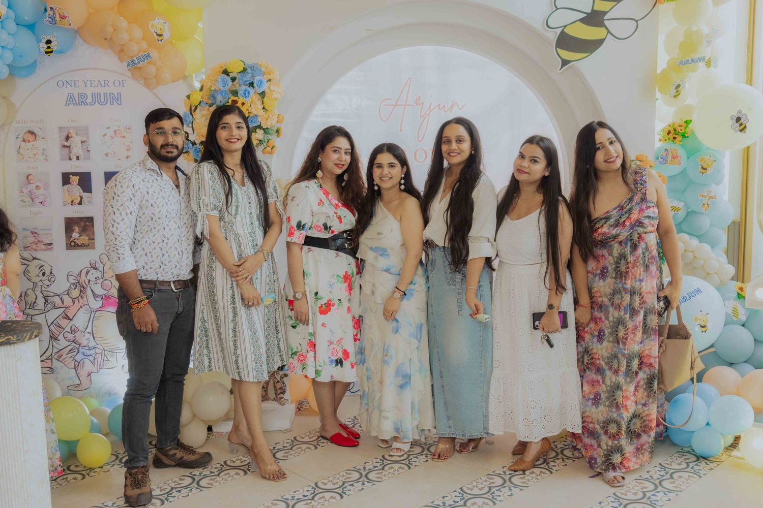 Group of seven friends posing together at a colorful baby shower party with balloon decorations.