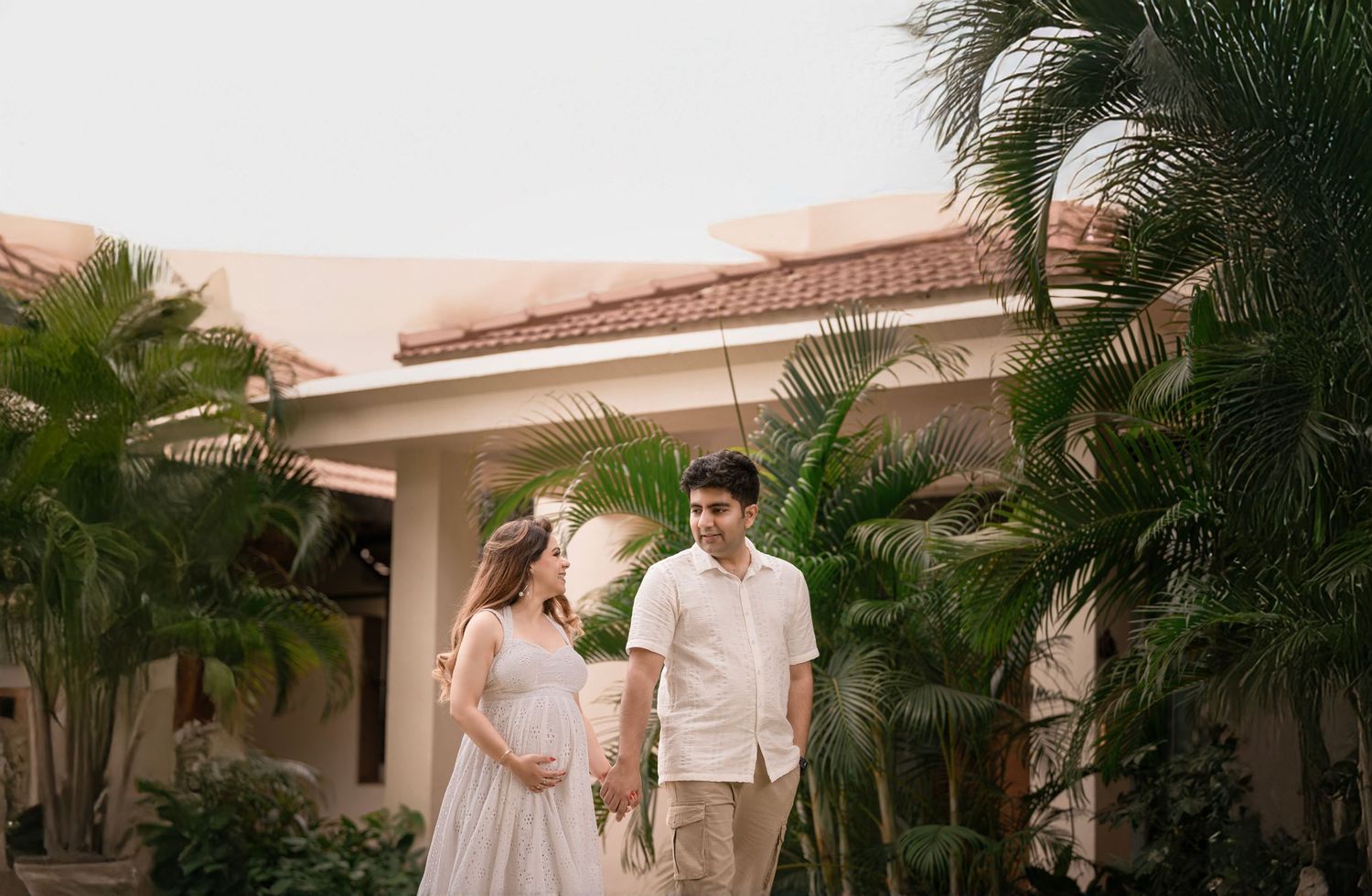 Pregnant couple holding hands outdoors near tropical palm trees and a beige villa during a maternity photoshoot.
