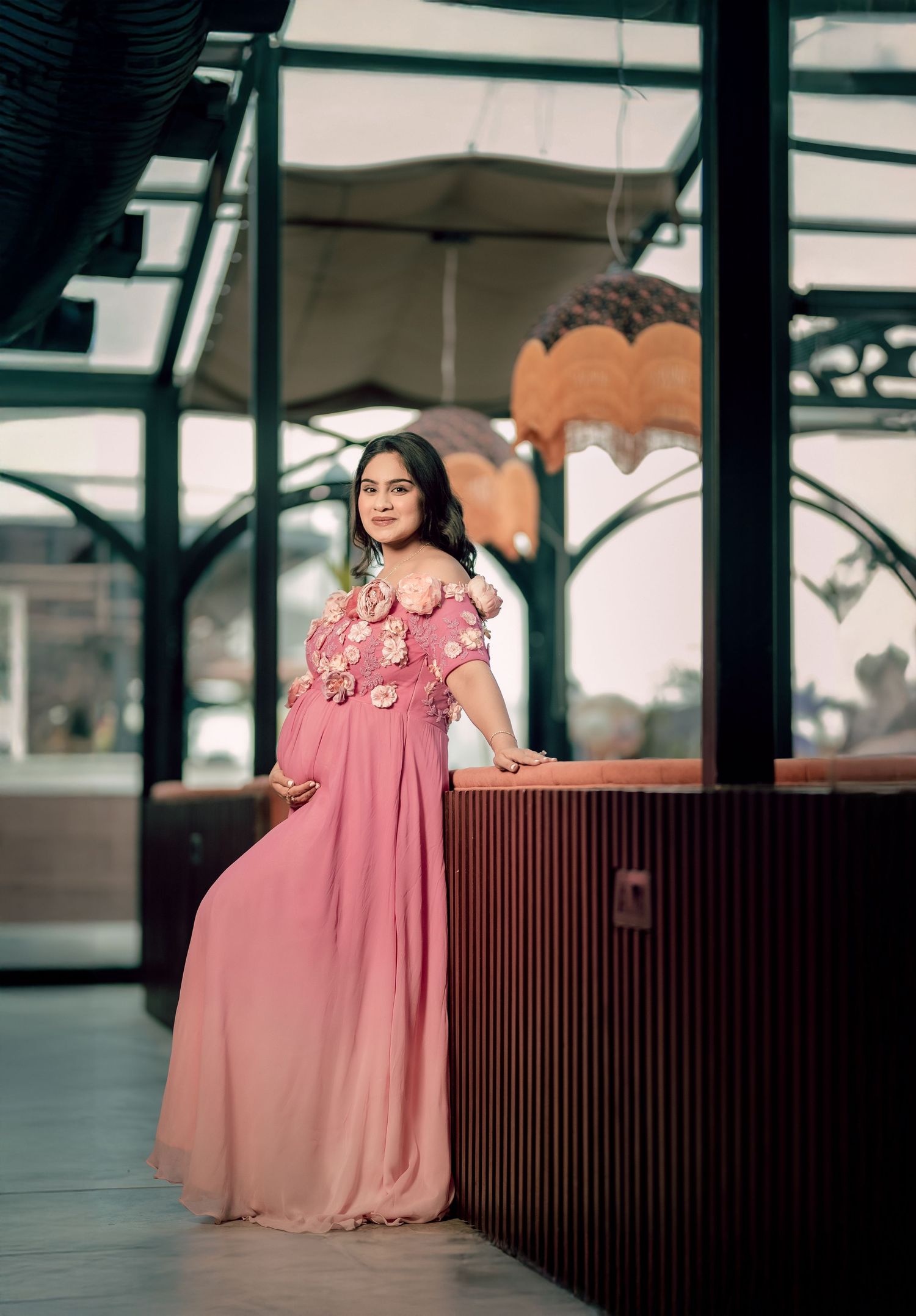 Woman in pink floral off-shoulder maxi gown posing elegantly at a wooden bar counter in a modern venue.