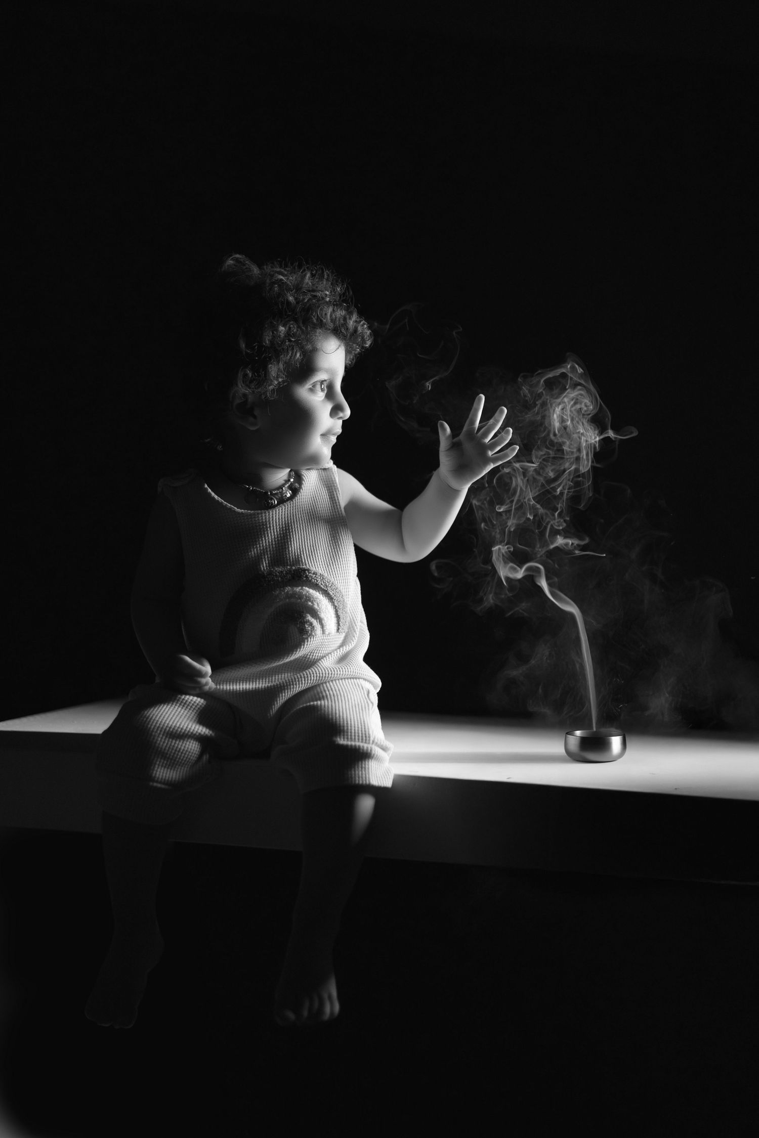 Black and white photo of a curious child reaching toward rising incense smoke on a dark background.
