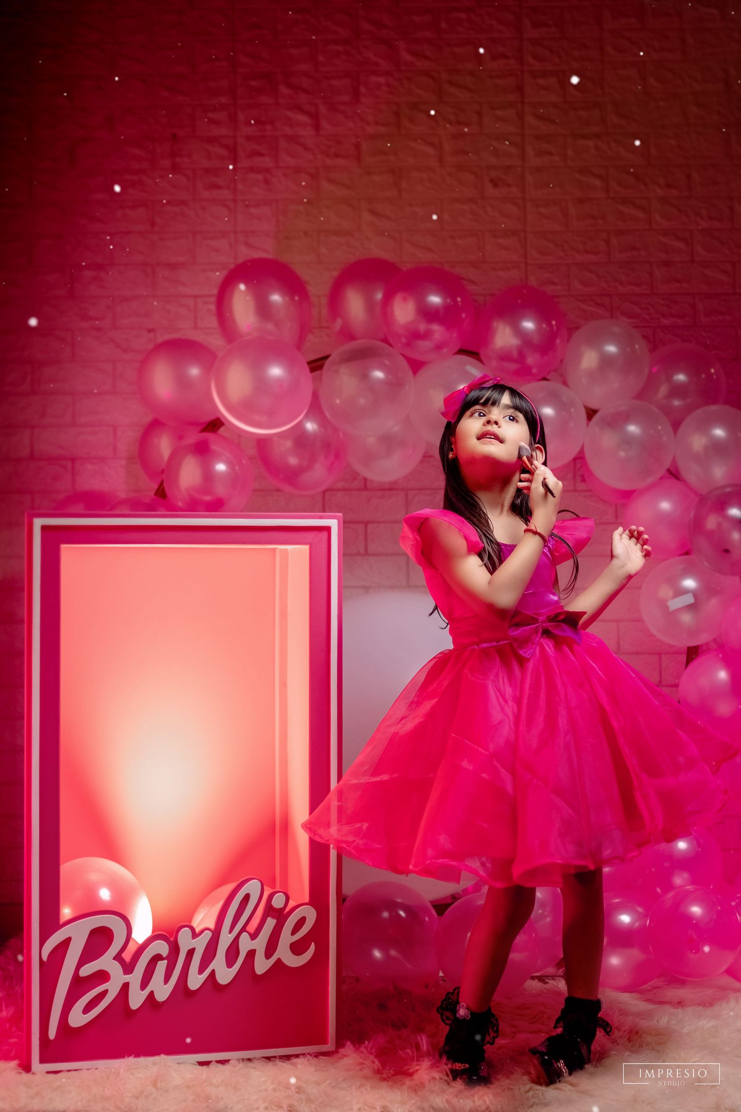 A young girl in a hot pink Barbie-themed dress poses beside a Barbie logo sign surrounded by pink balloons.