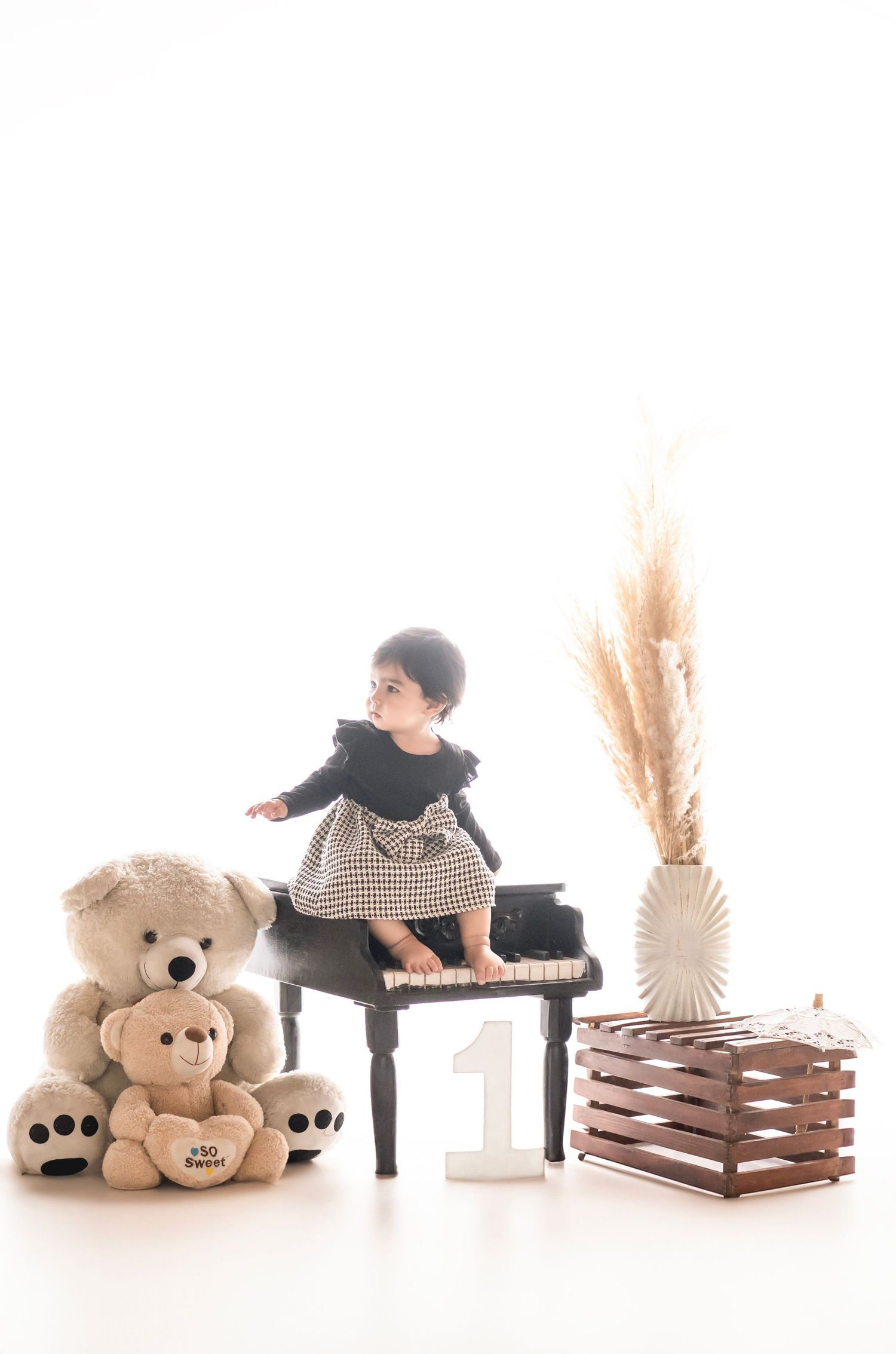 Baby girl in black dress sits on mini piano beside teddy bears and number 1, celebrating her first birthday.