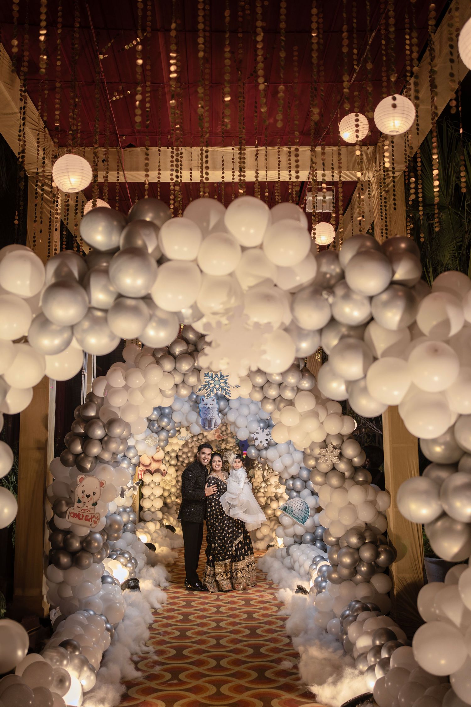 Couple poses under elegant white and silver balloon arch with hanging lanterns at a romantic wedding celebration.