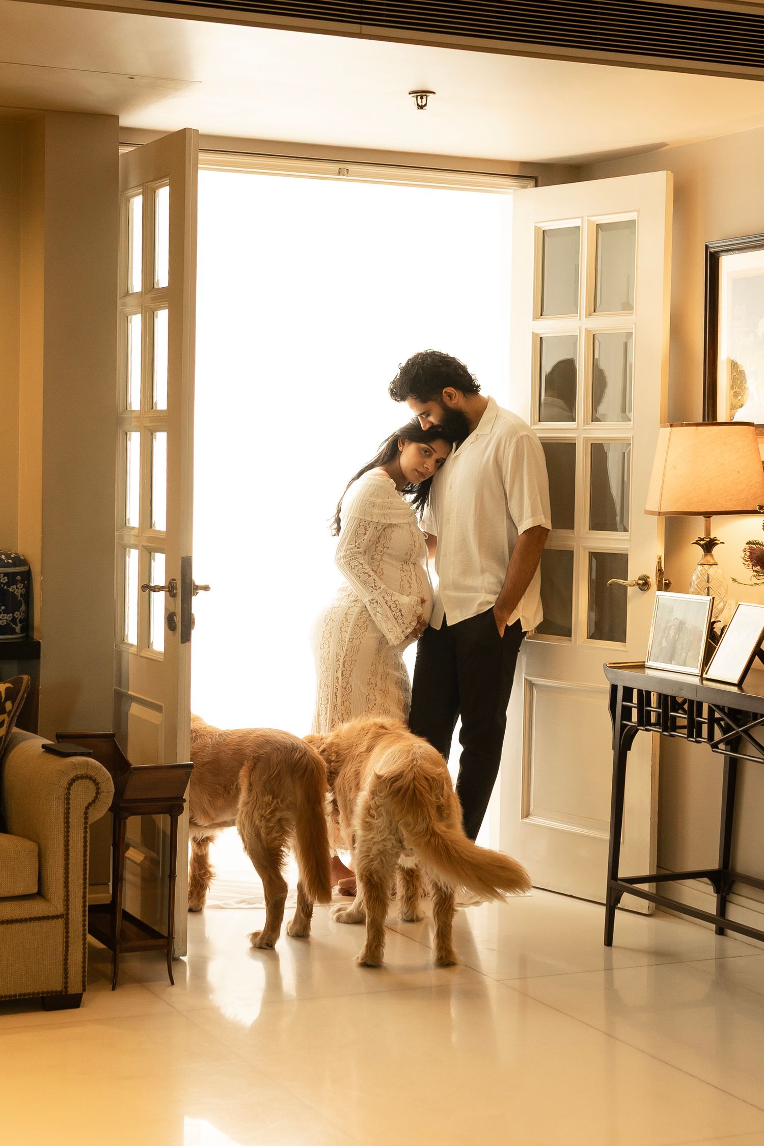 A romantic couple stands in a doorway with two golden retrievers in a warmly lit interior space for indoor maternity shoot.