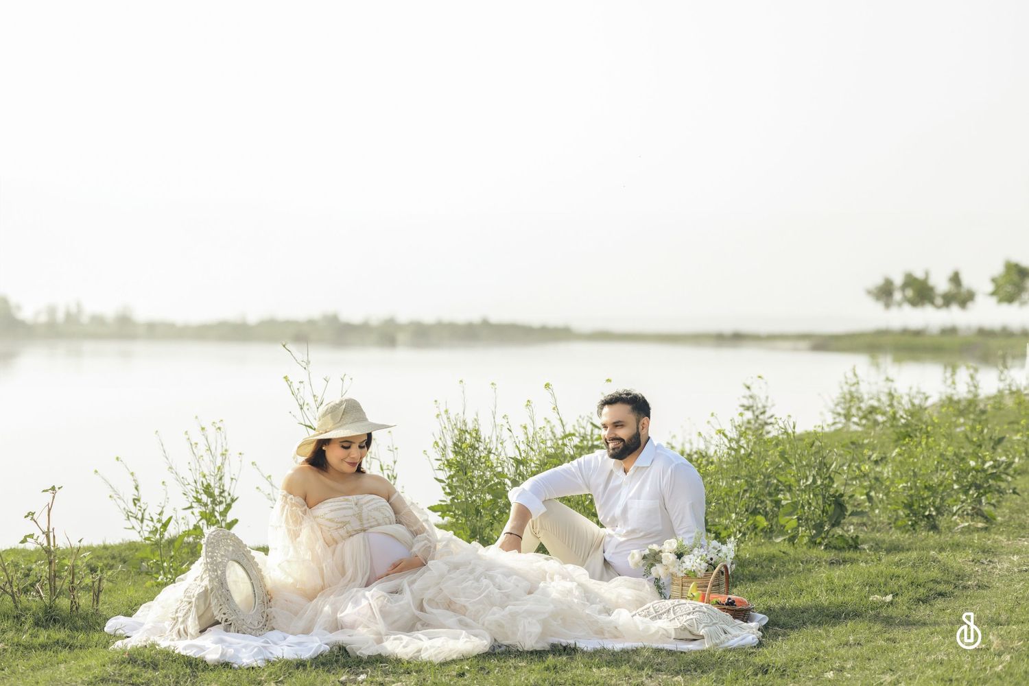 A couple enjoys a romantic lakeside picnic on a grassy shore with water and trees in the background for their maternity photoshoot.