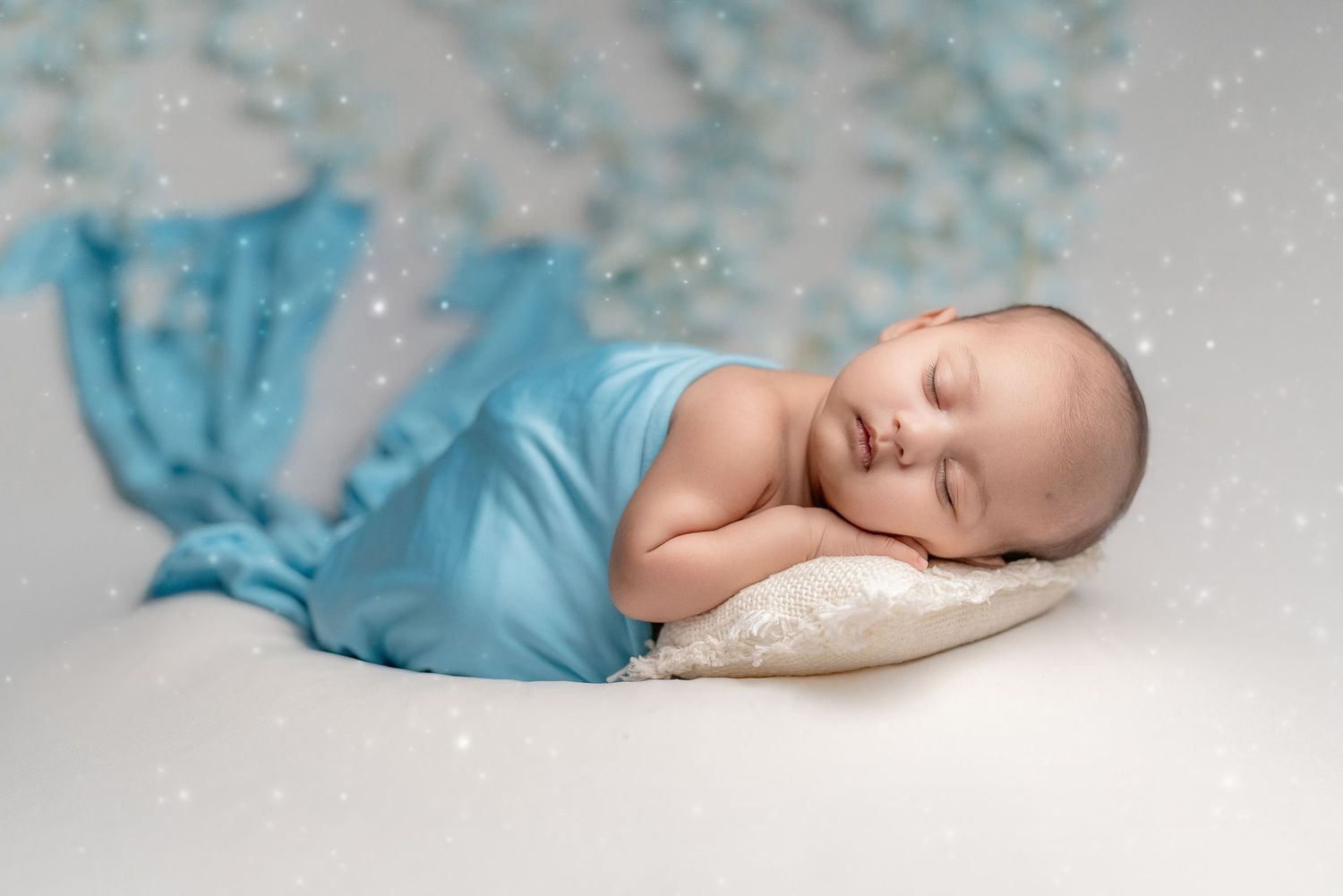 A sleeping newborn baby wrapped in soft white and turquoise fabric against a neutral background.