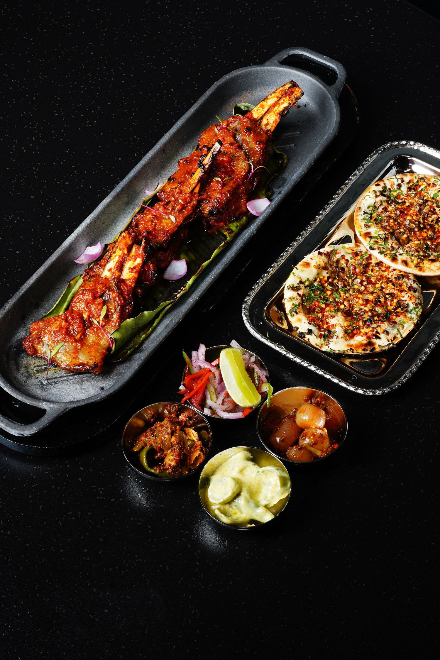 Grilled tandoori chicken skewers with naan bread and garnishes served on black plates against dark background.