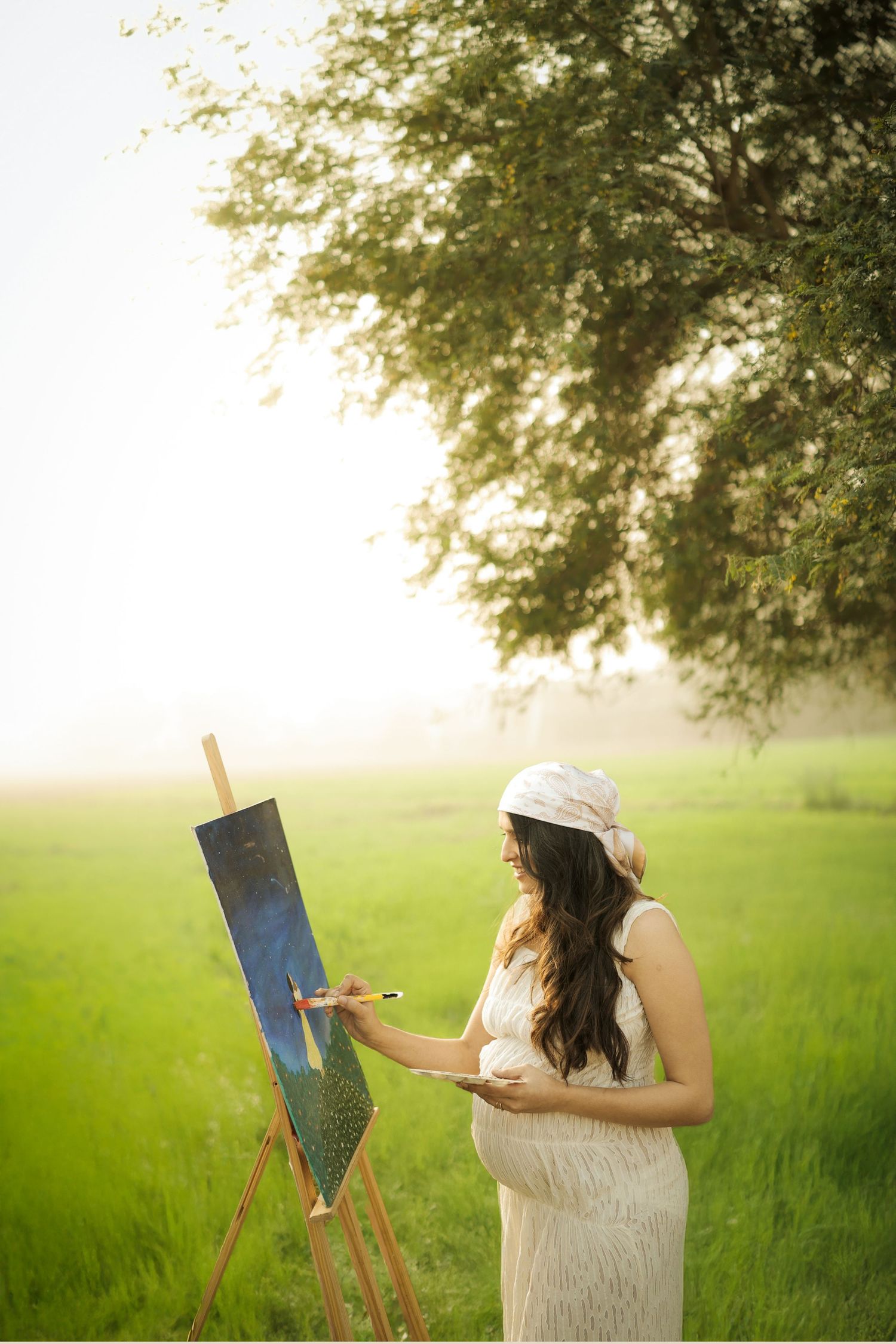Artist in white dress painting on canvas outdoors in a sunlit green meadow during golden hour.