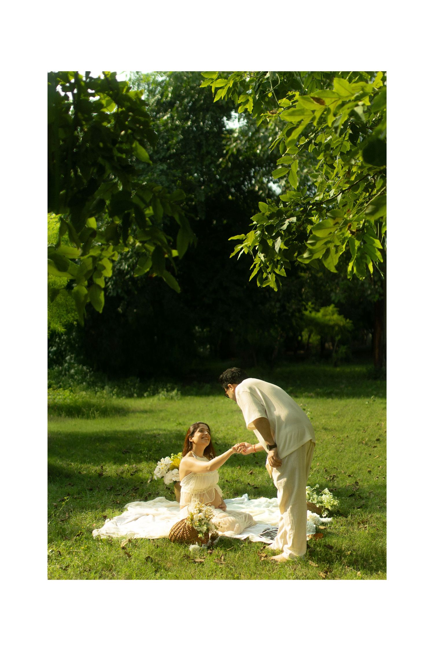 A couple in wedding attire lay on a white blanket in a sunny garden setting during a romantic photoshoot.