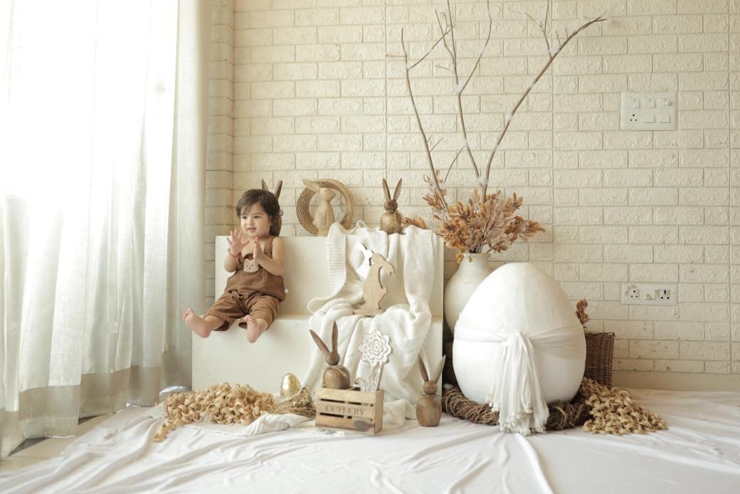A toddler sits beside Easter decorations including a large white egg, bunny figurines, and dried flowers in a bright white room.