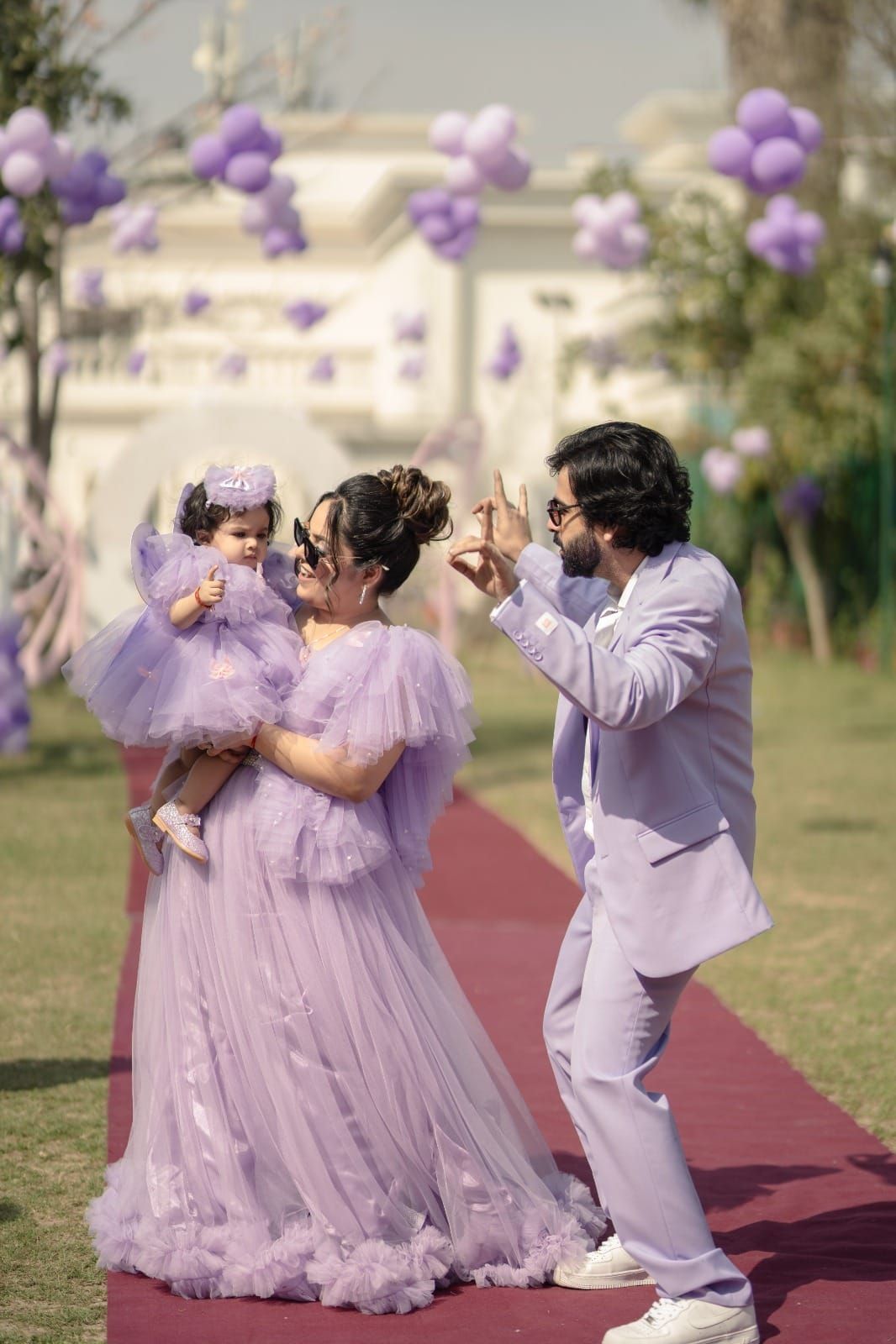 Family in matching lavender outfits celebrating outdoors with purple balloon decorations on a red carpet.