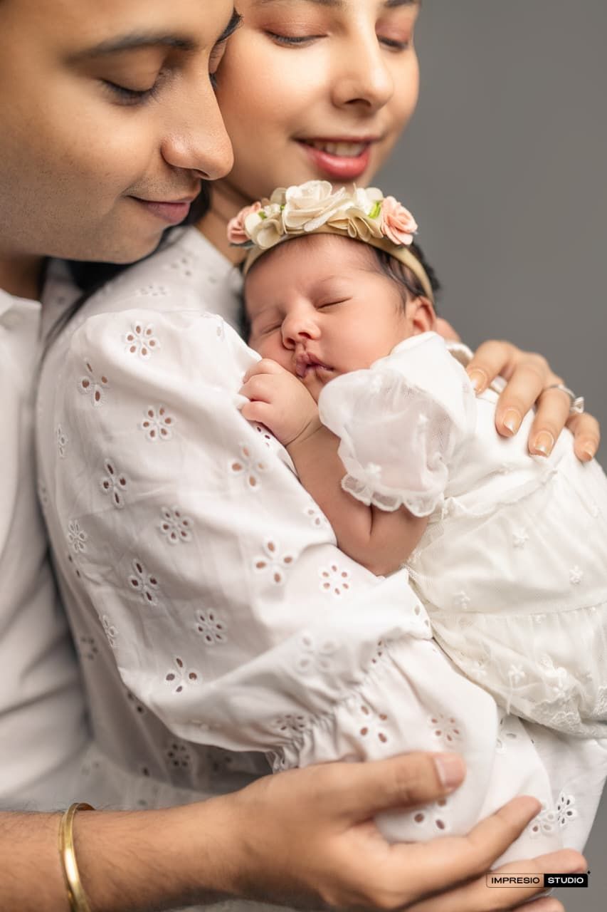 New parents tenderly hold their sleeping newborn baby girl in a white dress and floral headband.