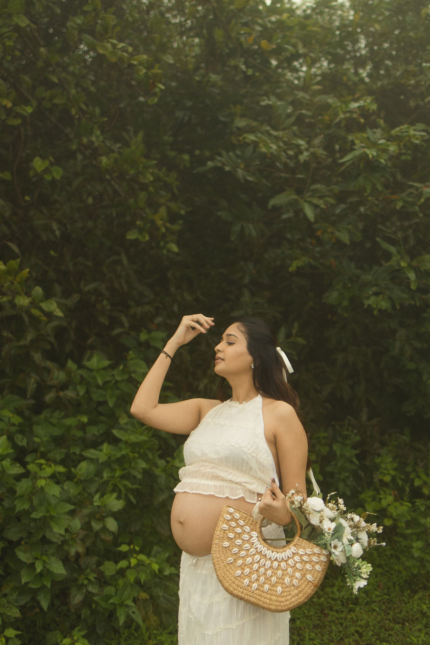 Pregnant woman in white dress holding wicker bag with flowers, posing outdoors among lush green foliage.
