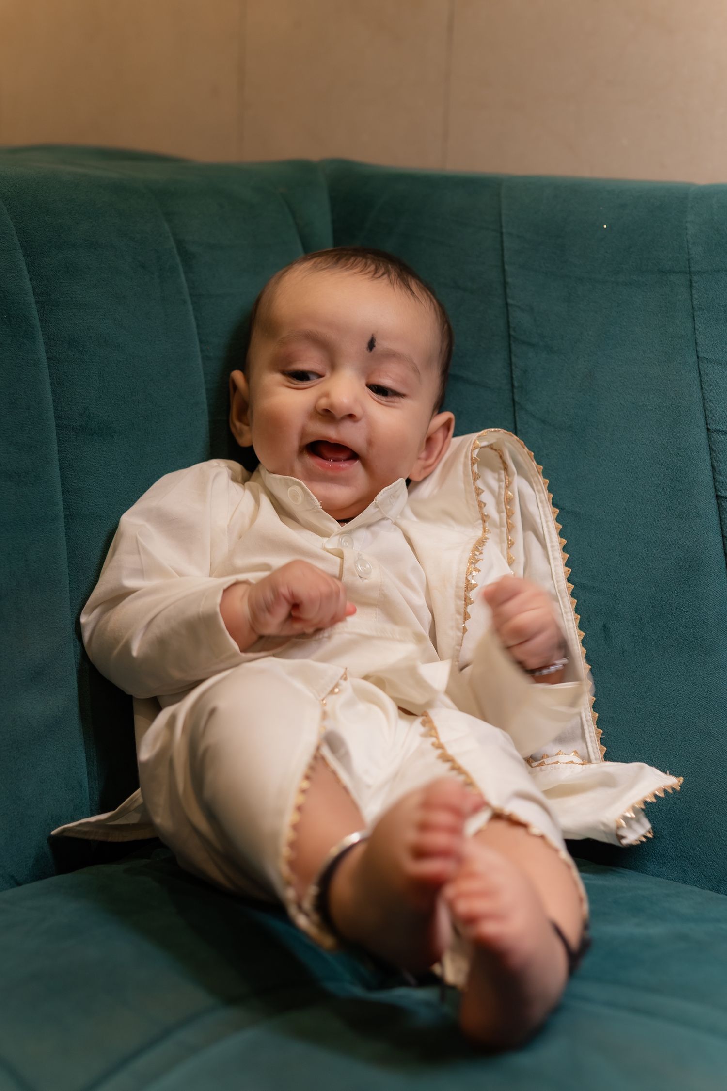 Baby in a cream traditional outfit with gold trim and a black dot on the forehead sits on a teal velvet sofa, looking down at their hands.