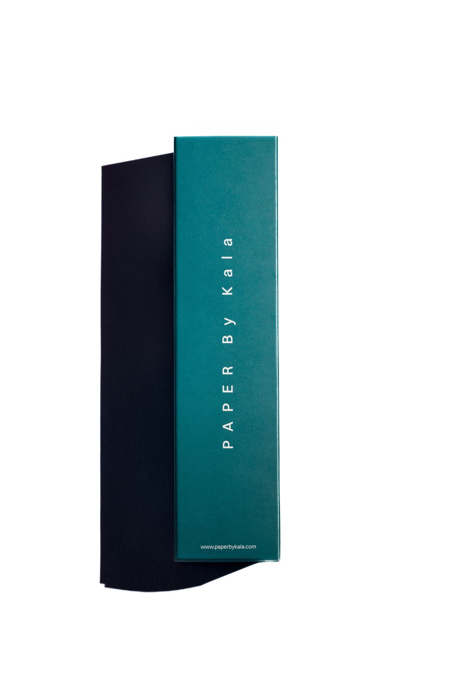 Teal green PAPER By Kala box with white vertical lettering, standing against a dark navy notebook or folder on a white background.