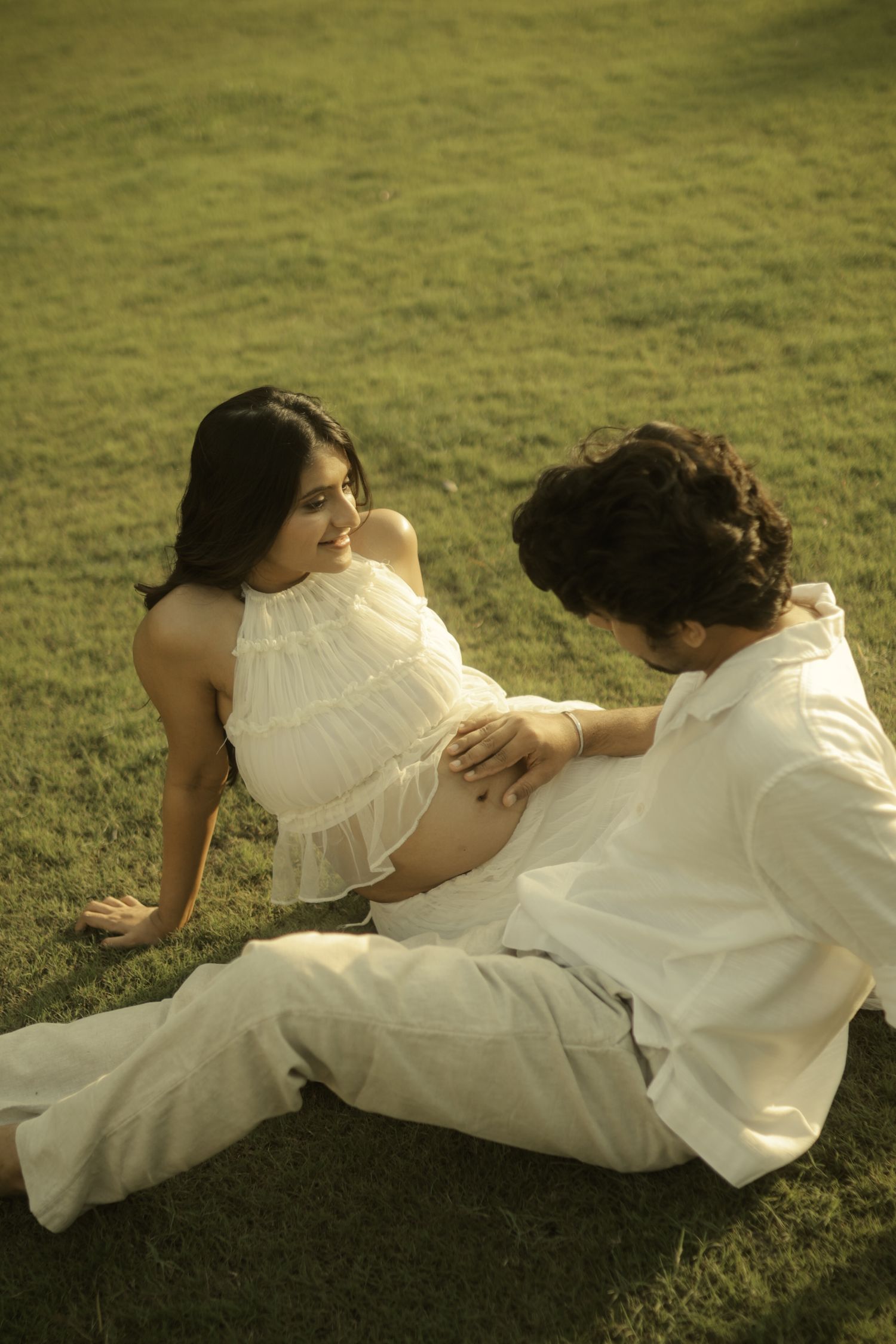 Pregnant woman in white dress sits on grass with partner touching her baby bump during outdoor maternity photoshoot.