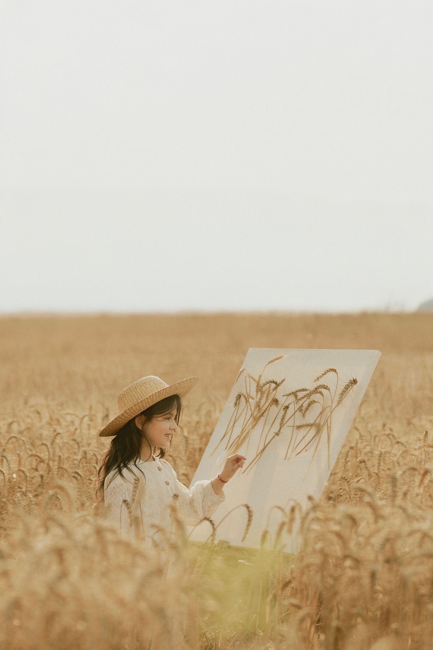 A young woman in a straw hat paints wheat stalks on canvas while standing in a golden wheat field.