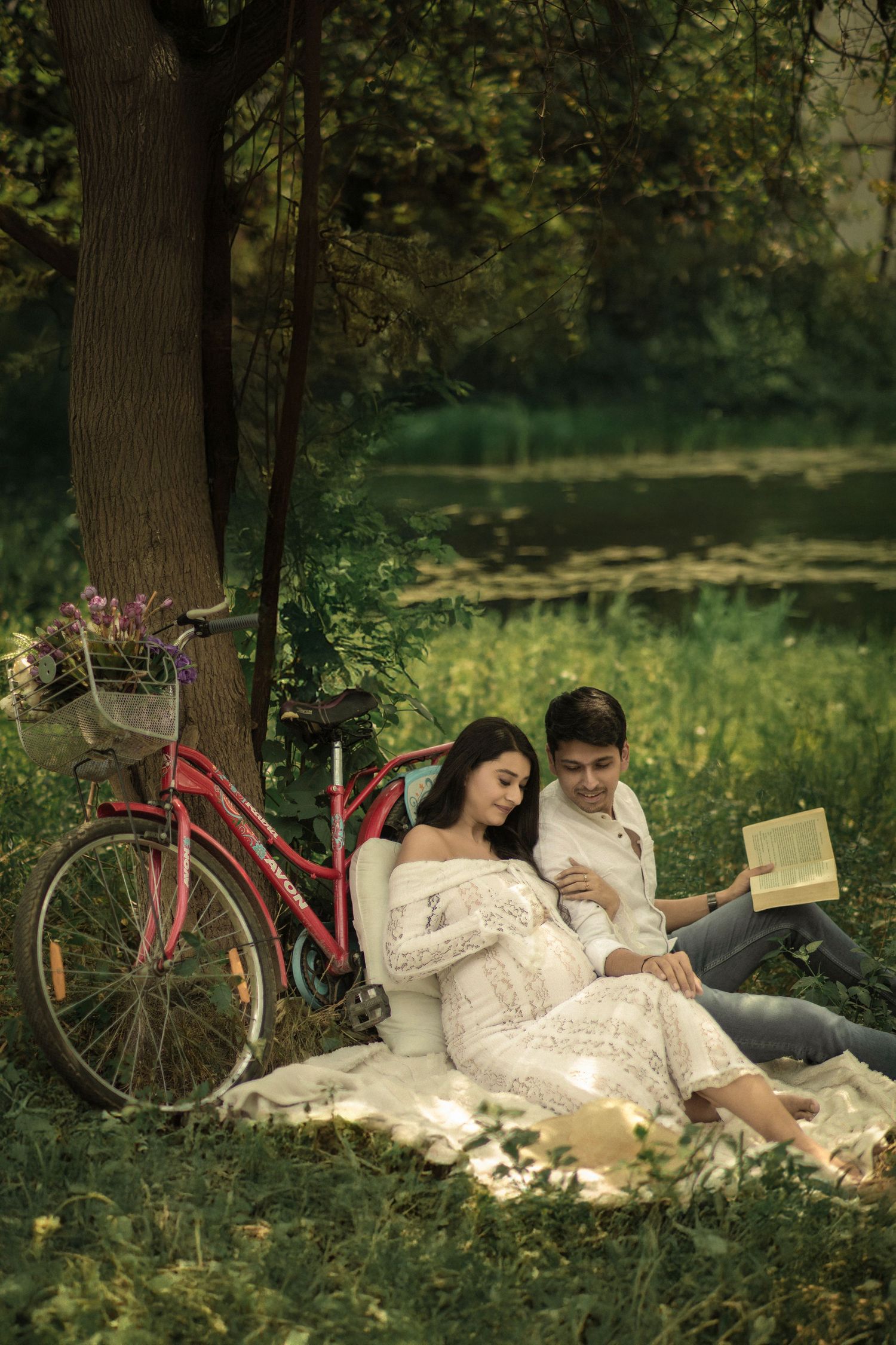 Romantic couple reading a book together on a blanket under a tree by a pond, with a vintage pink bicycle nearby.