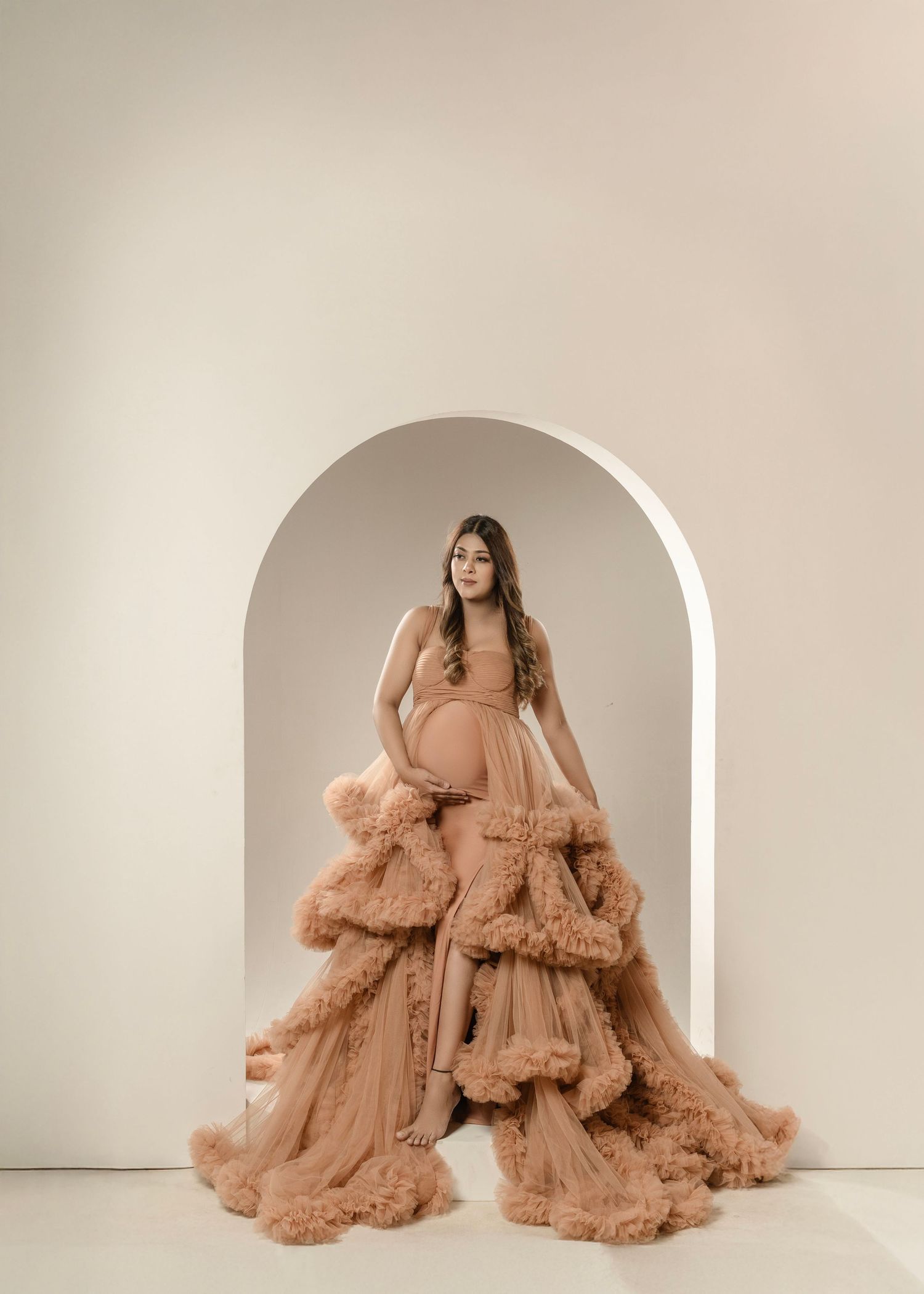 Studio maternity photograph with luxurious ruffled brown tulle gown in arched backdrop.