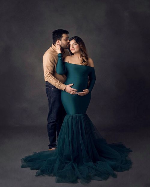Elegant maternity portrait with couple in teal mermaid gown against dark studio backdrop.