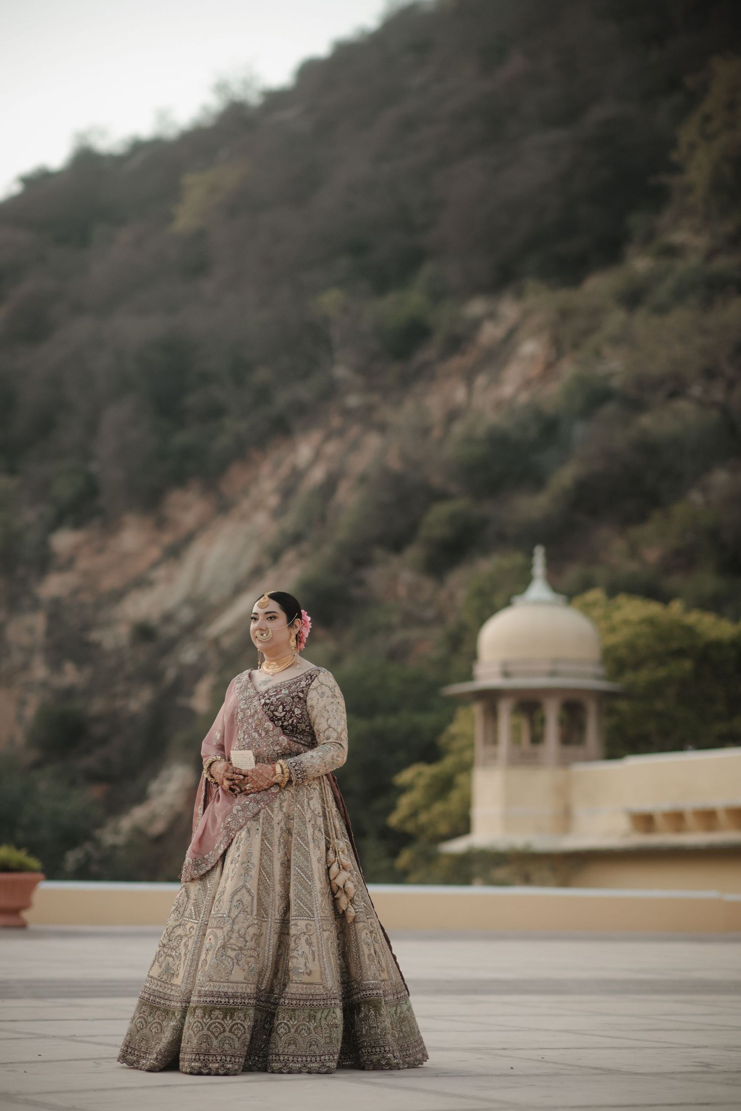 Elegant bridal portrait featuring ornate lehenga against mountainous backdrop with domed architecture.
