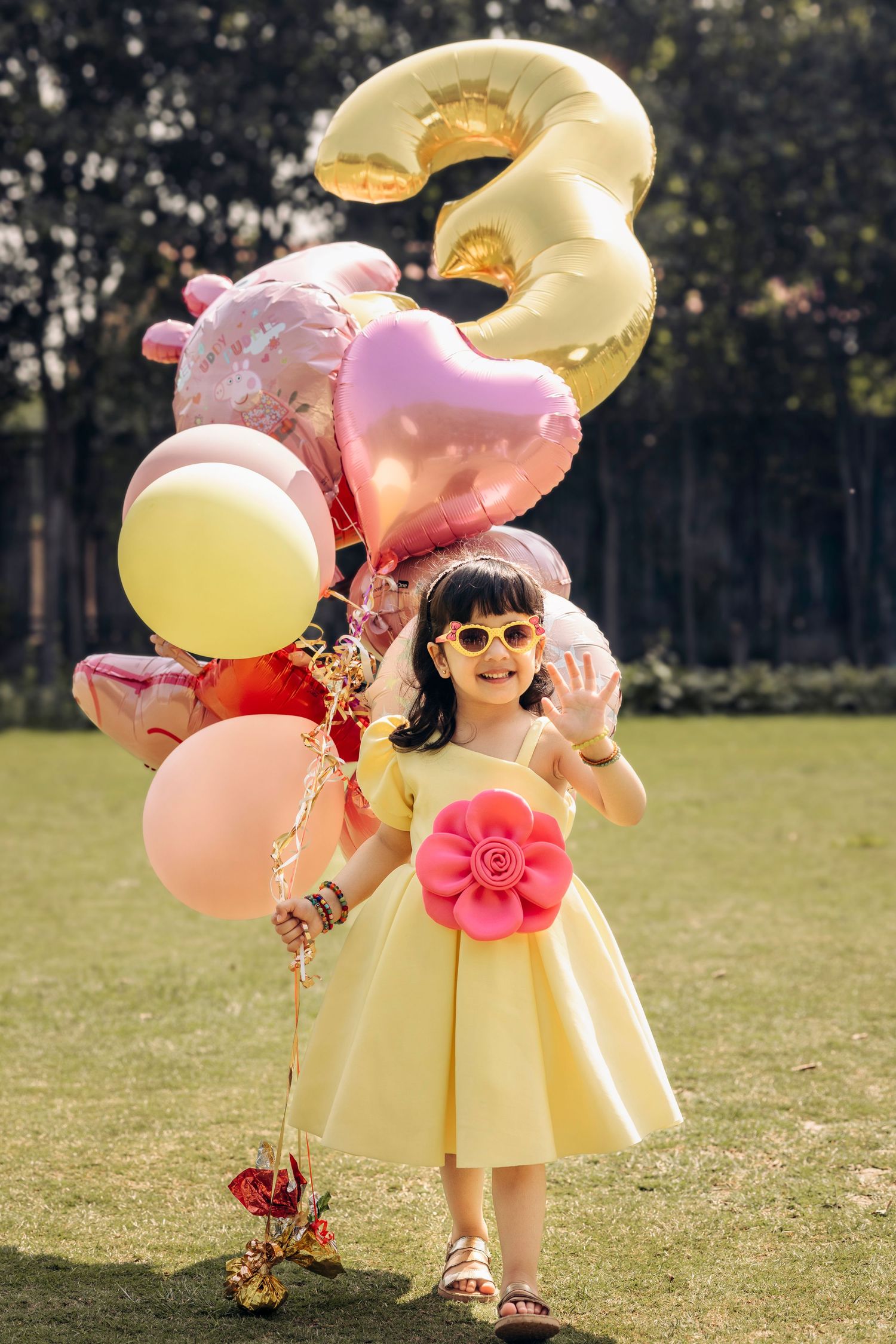 A child in a yellow dress with pink bow holds colorful balloons including gold number 3 while standing on grass outdoors.