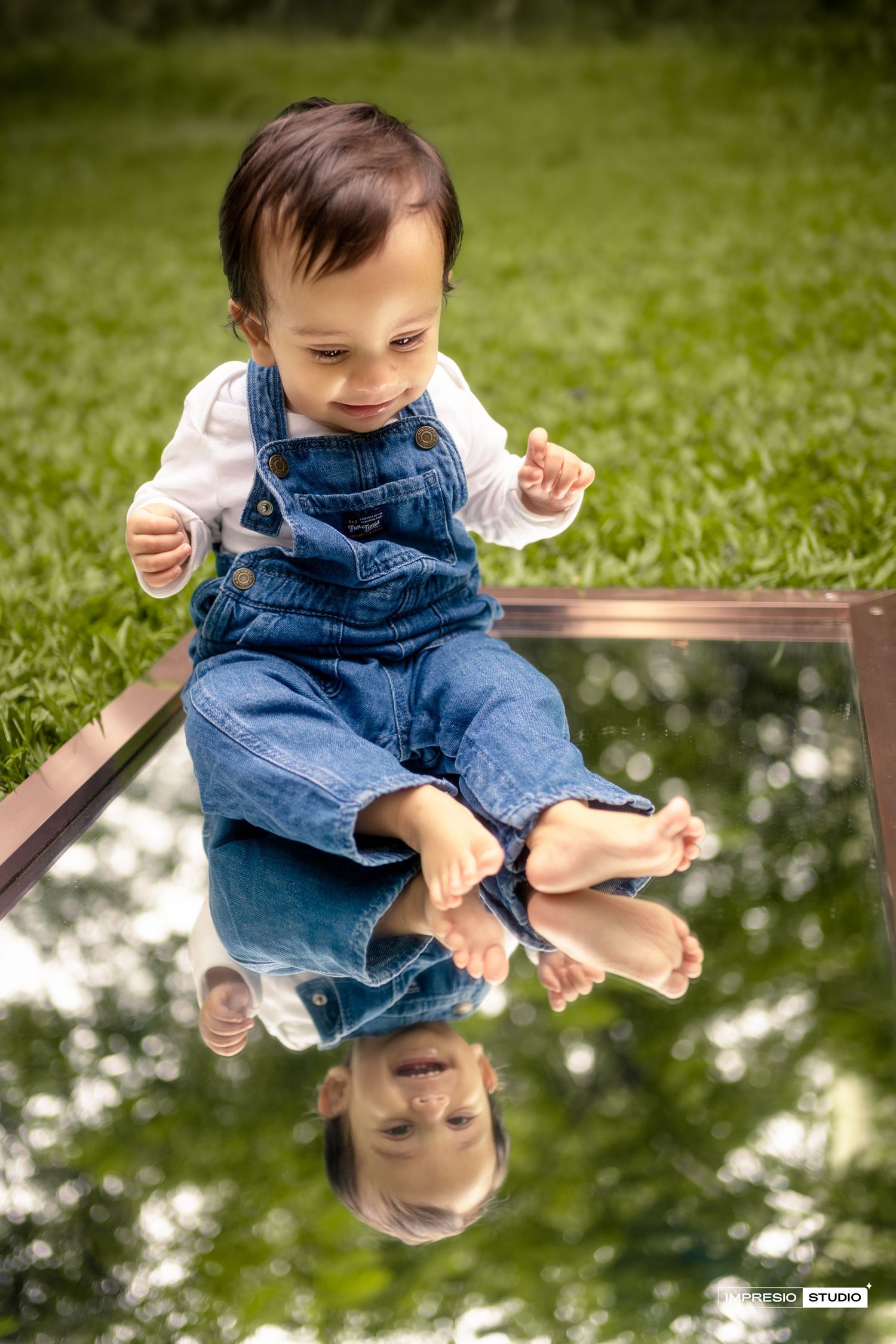 A toddler in denim overalls sits on a mirrored surface outdoors surrounded by grass and white flowers.