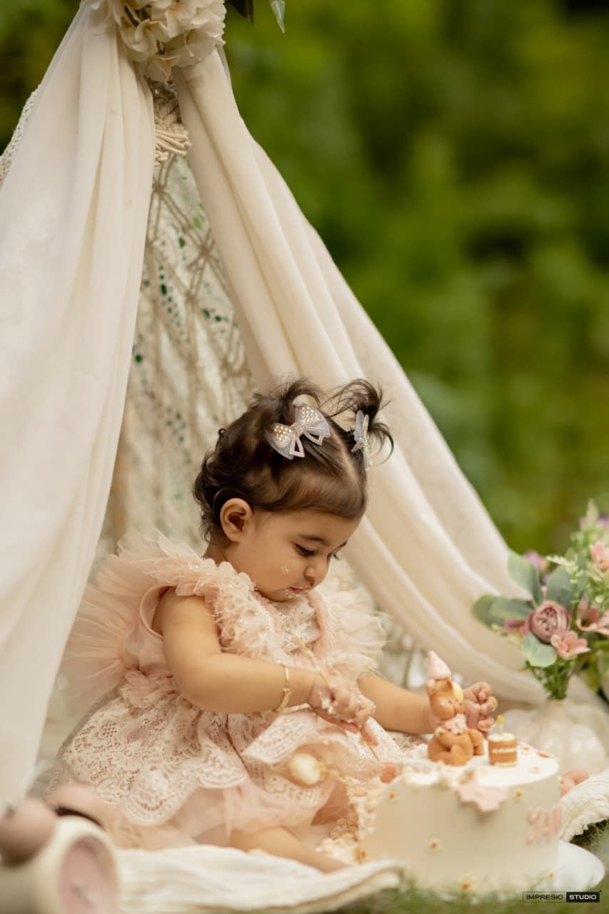 Toddler girl in pink lace dress plays with cake inside a boho teepee tent outdoors.