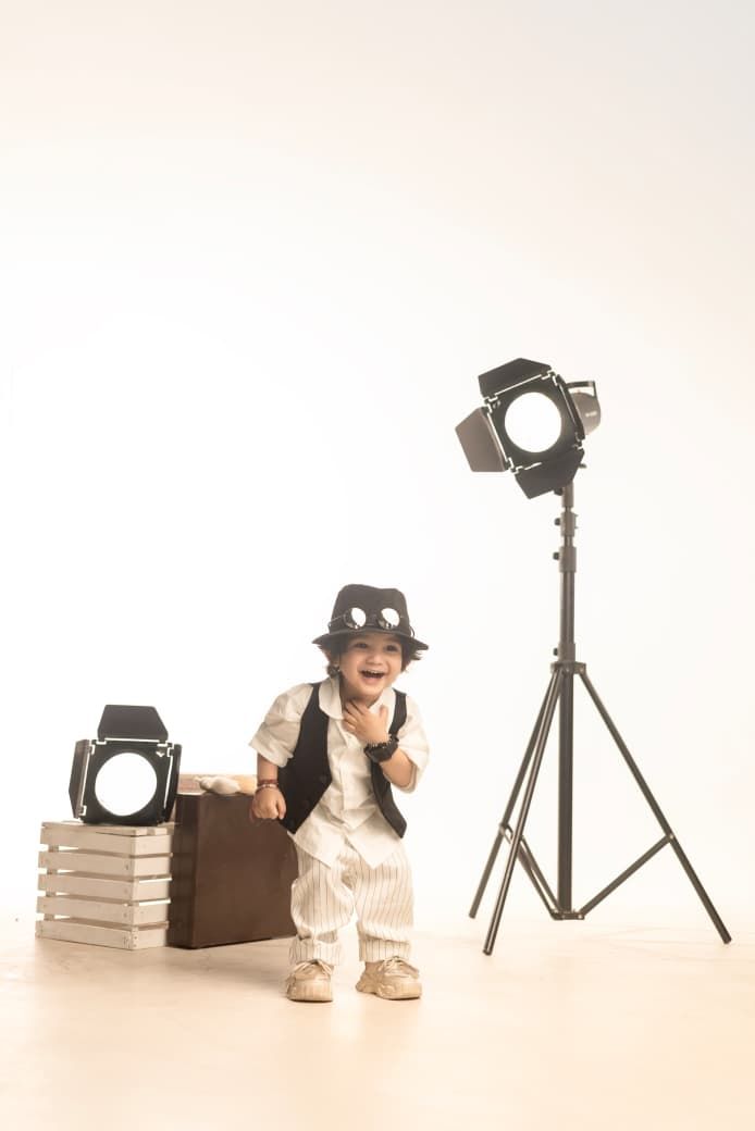 A cheerful toddler dressed in a white suit and black hat poses with vintage suitcase and studio lights on white background.