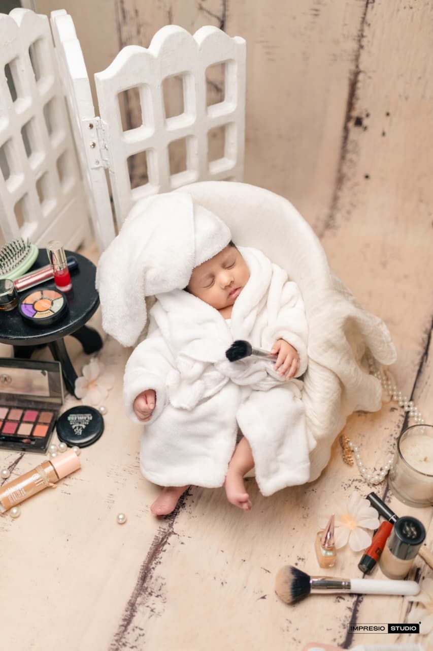 Sleeping newborn baby in white robe and towel turban sits in tiny chair surrounded by makeup and beauty products.