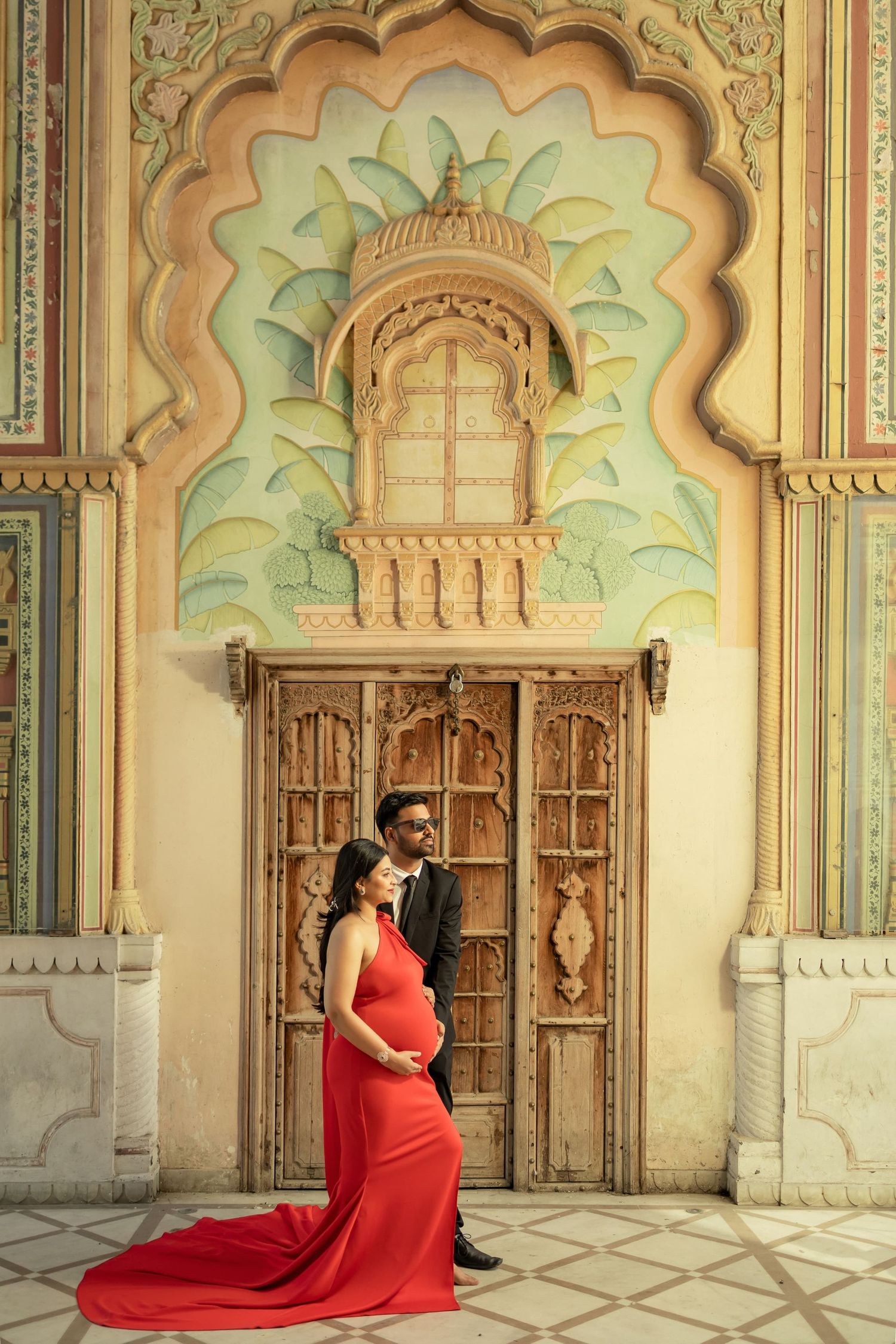 Couple stands in front of an ornate archway for maternity photoshoot with traditional Indian architectural details and decorative carvings.