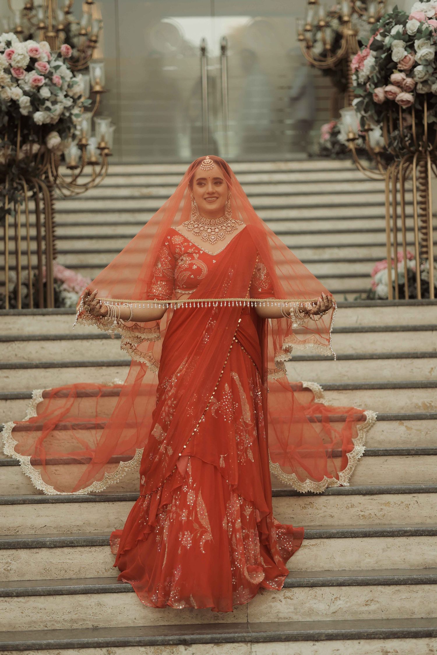 Bride in red embroidered saree with veil on marble stairs, adorned with gold jewelry and floral arrangements.