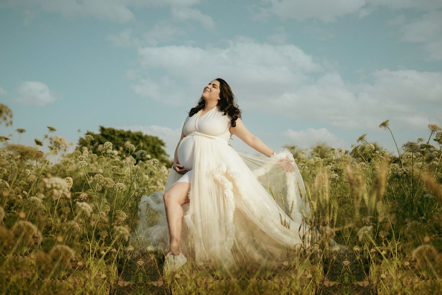 Beautiful maternity portraits in flowing white dress among wildflowers at sunset.