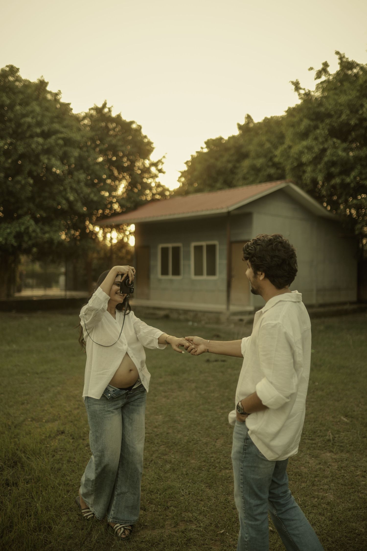 A pregnant couple holds hands outdoors at sunset, wearing white shirts and jeans, with a small house and trees in the background.