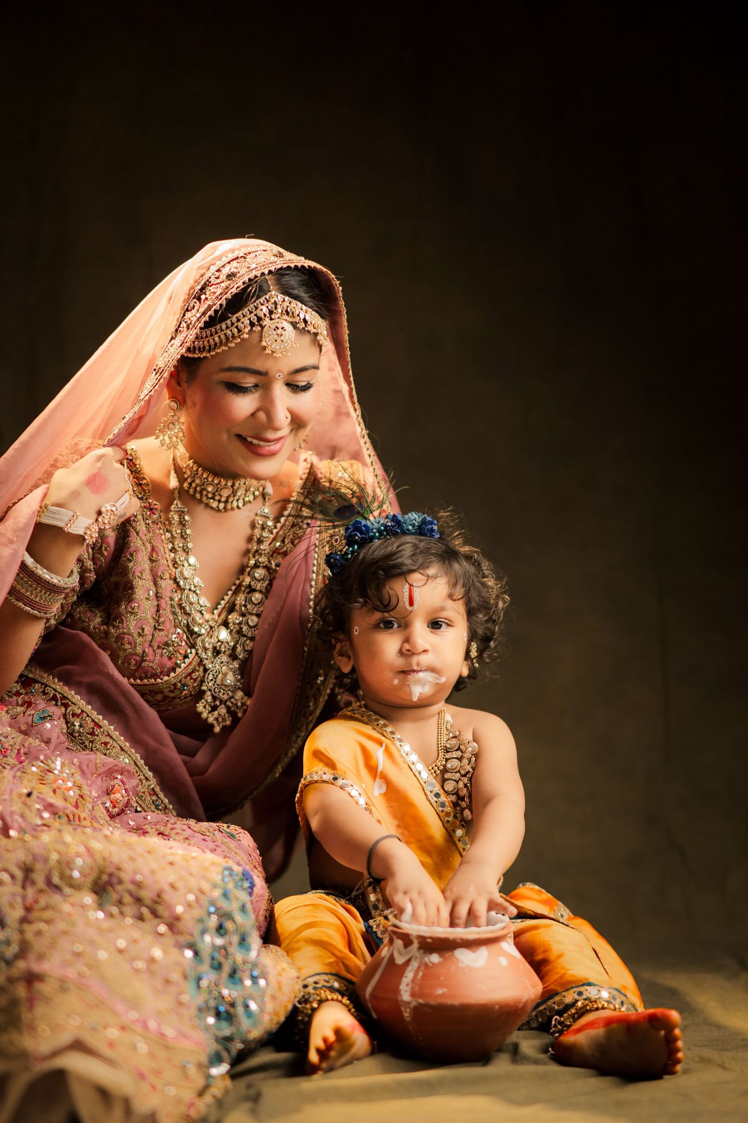 Indian woman in traditional bridal attire smiling at baby dressed as Lord Krishna playing with clay pot.