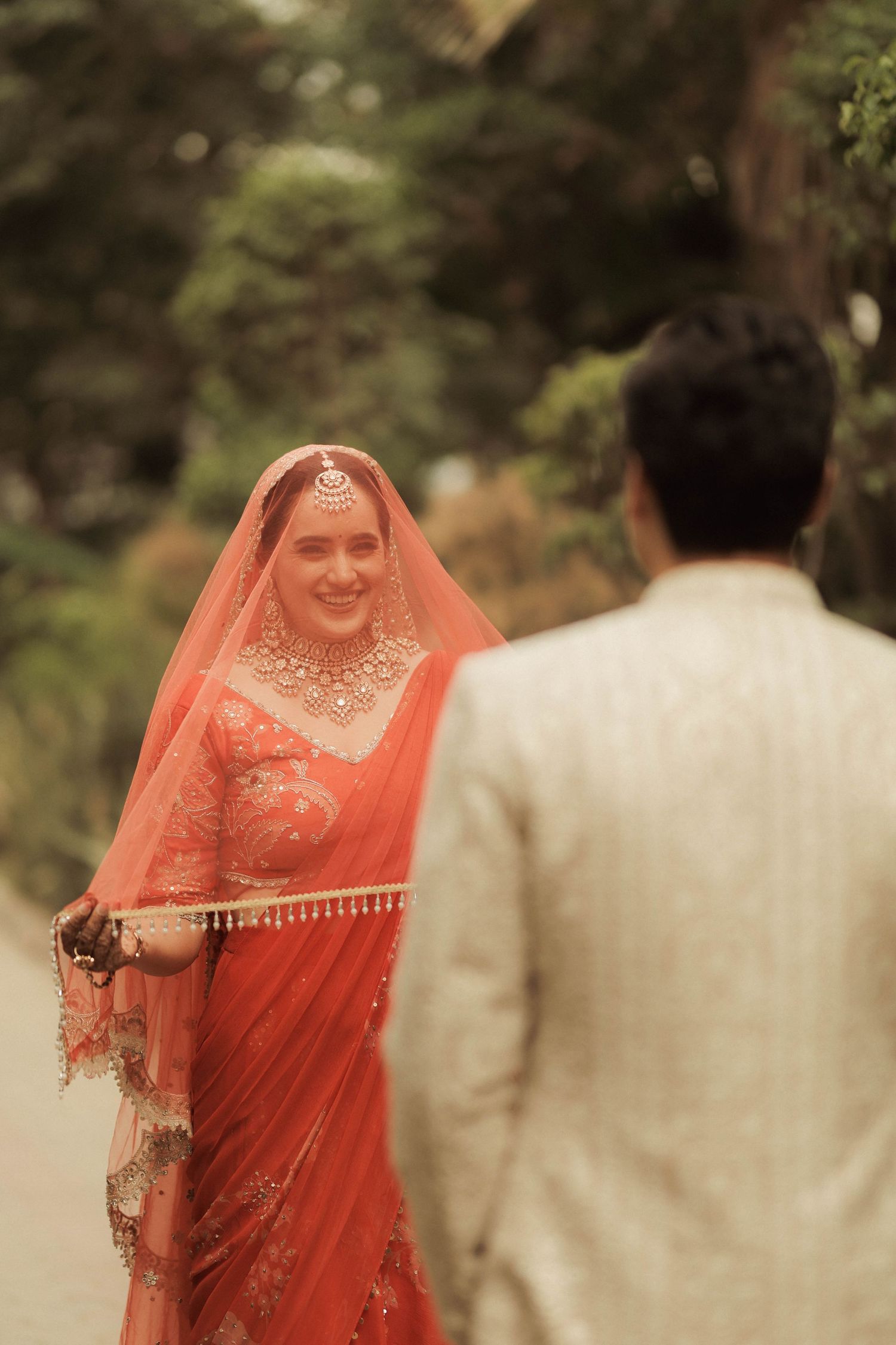 Indian bride in red saree smiling at groom in cream suit during traditional wedding ceremony outdoors.
