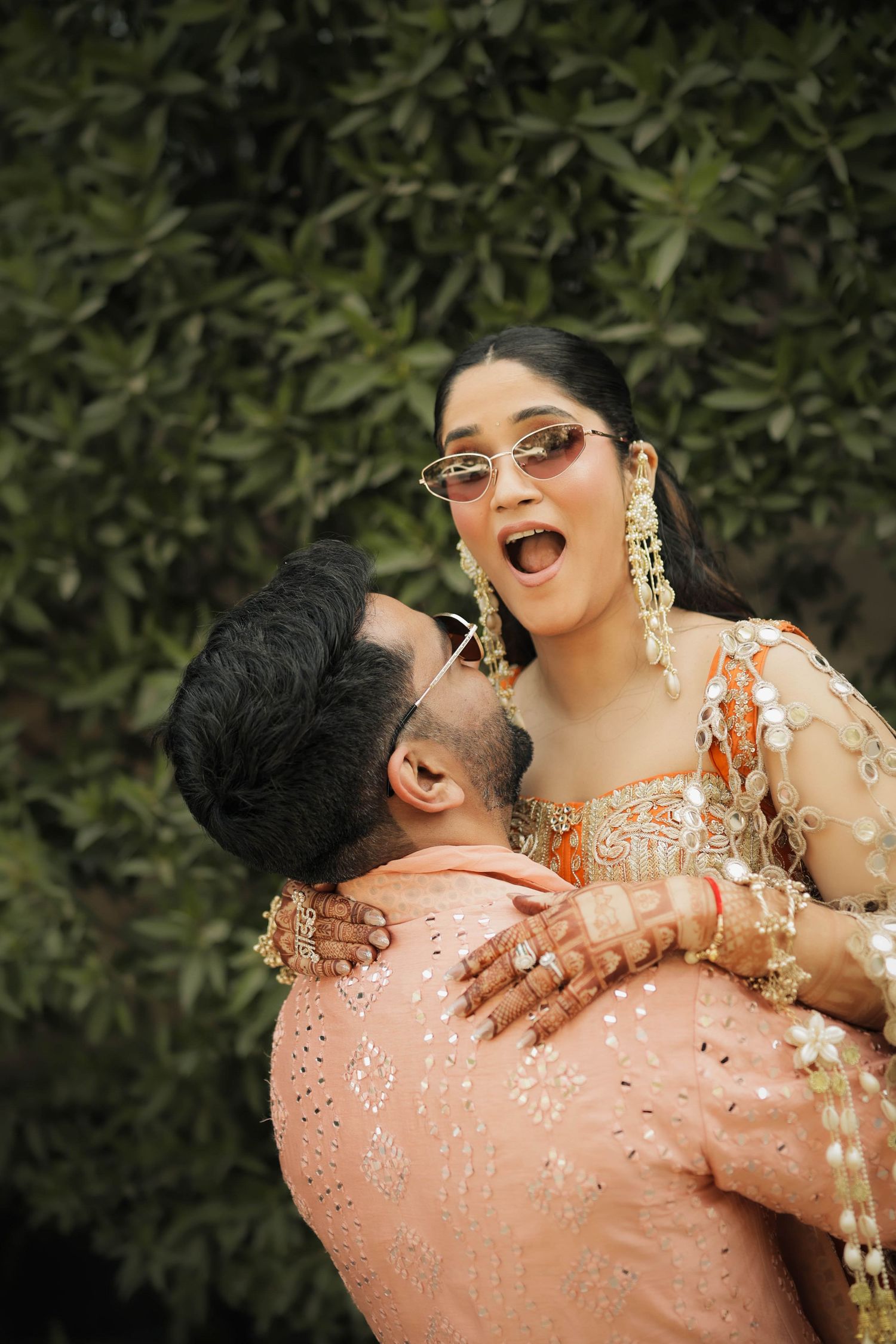 A joyful couple in traditional Indian wedding attire, man lifting woman who wears sunglasses, green foliage background.